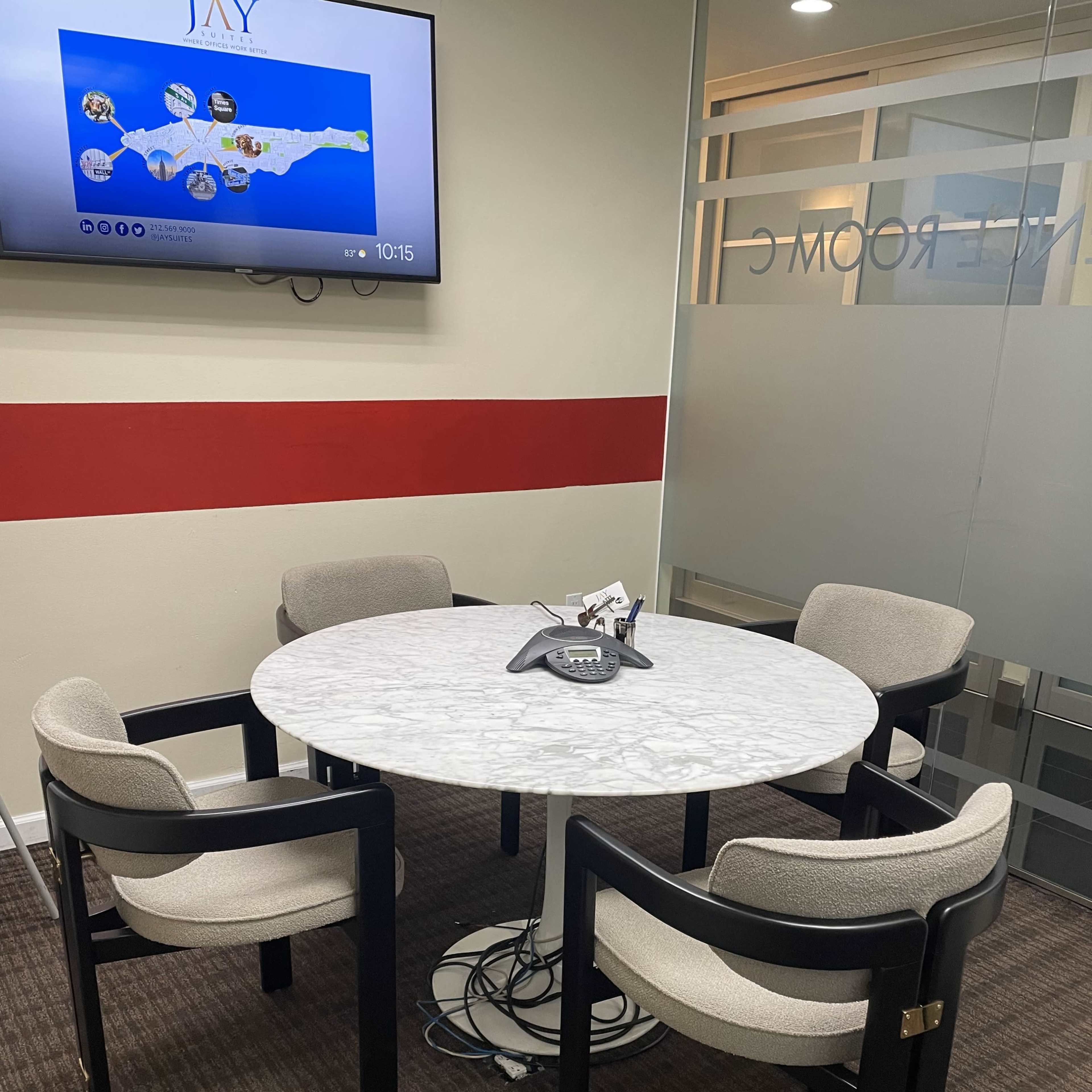 A round marble table with four cushioned chairs is positioned in a conference room with a television displaying a map.