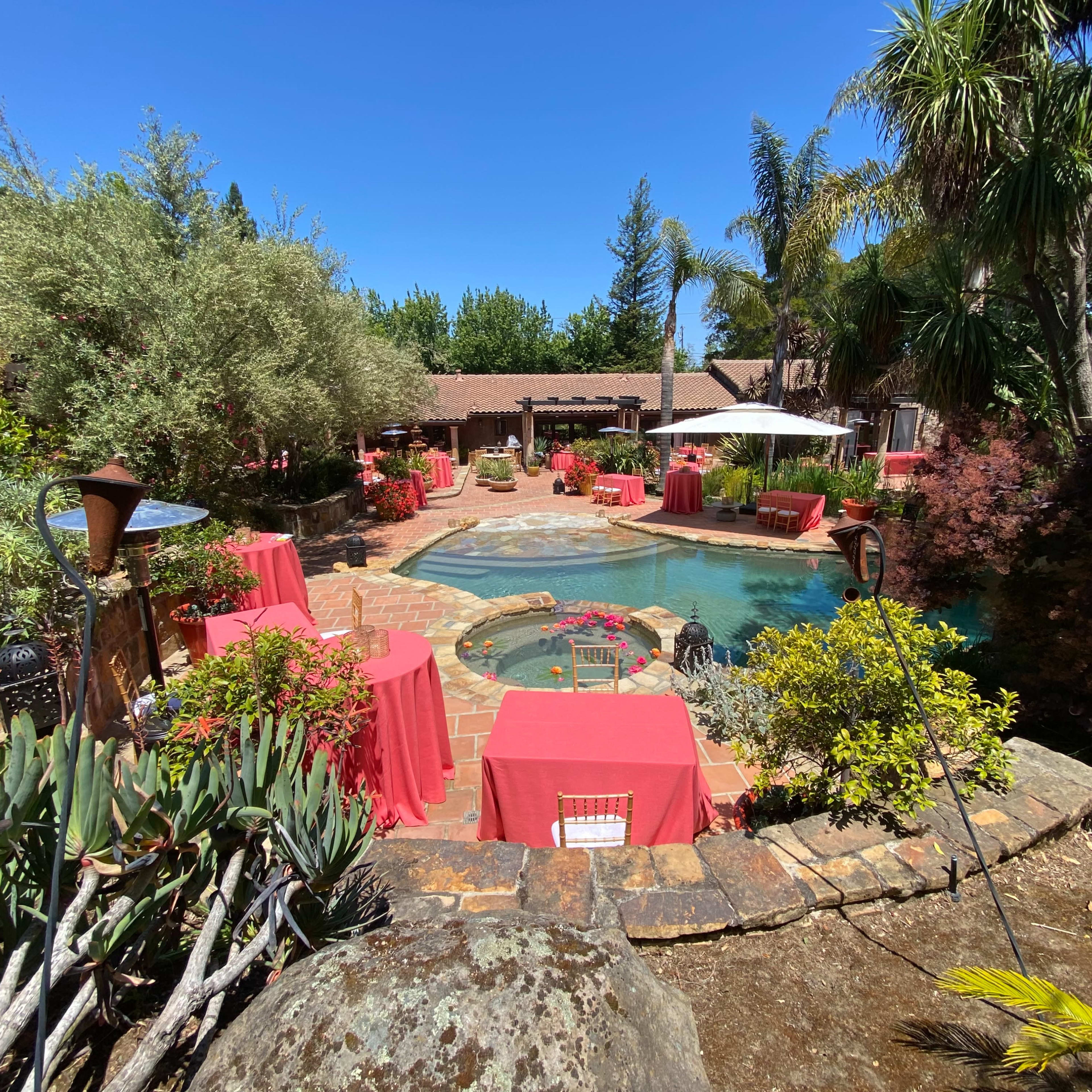A landscaped pool area surrounded by tables with red tablecloths and lush greenery.