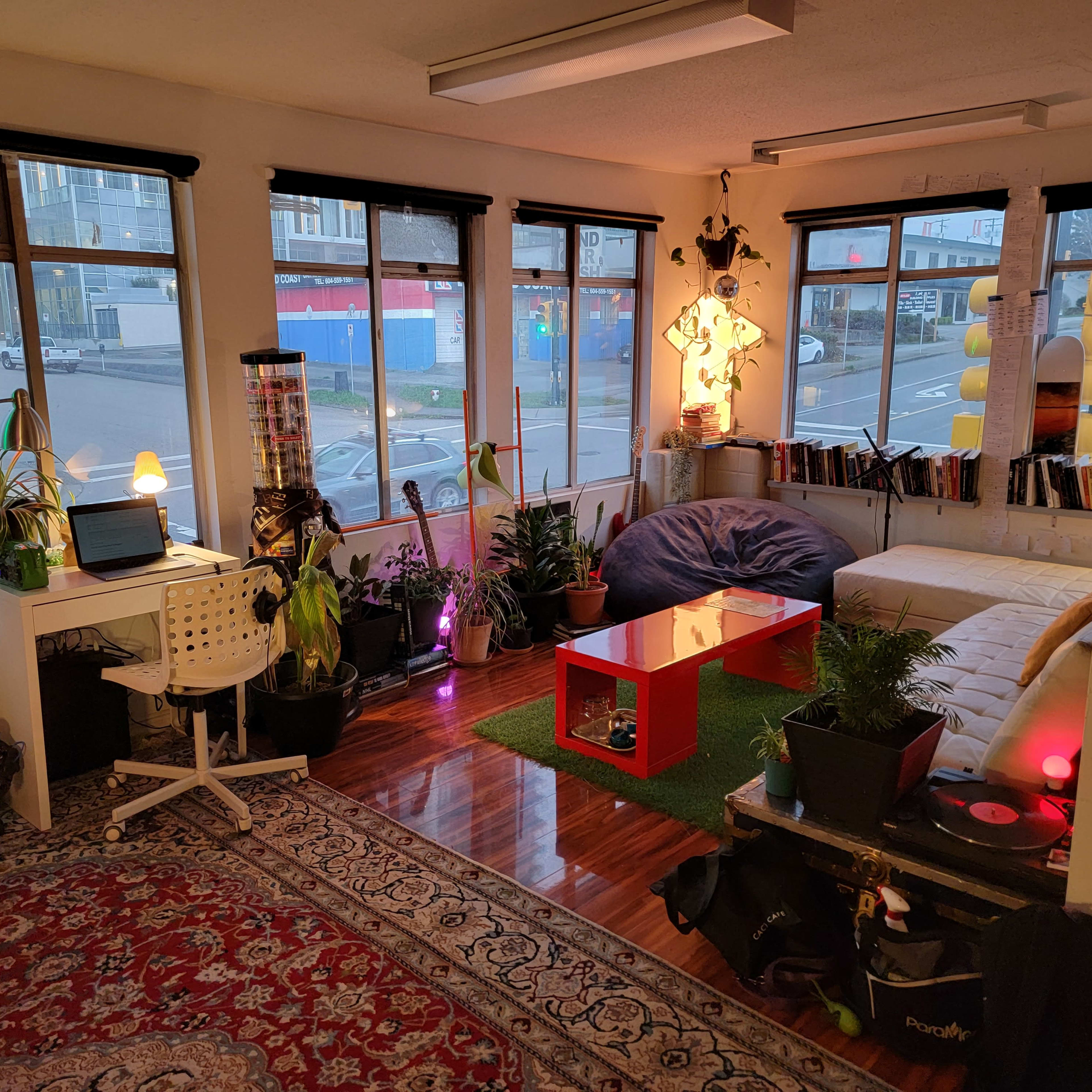 A cozy room with large windows, featuring a seating area with a white sofa, a low red coffee table, several plants, a dedicated workspace, and colorful lighting.