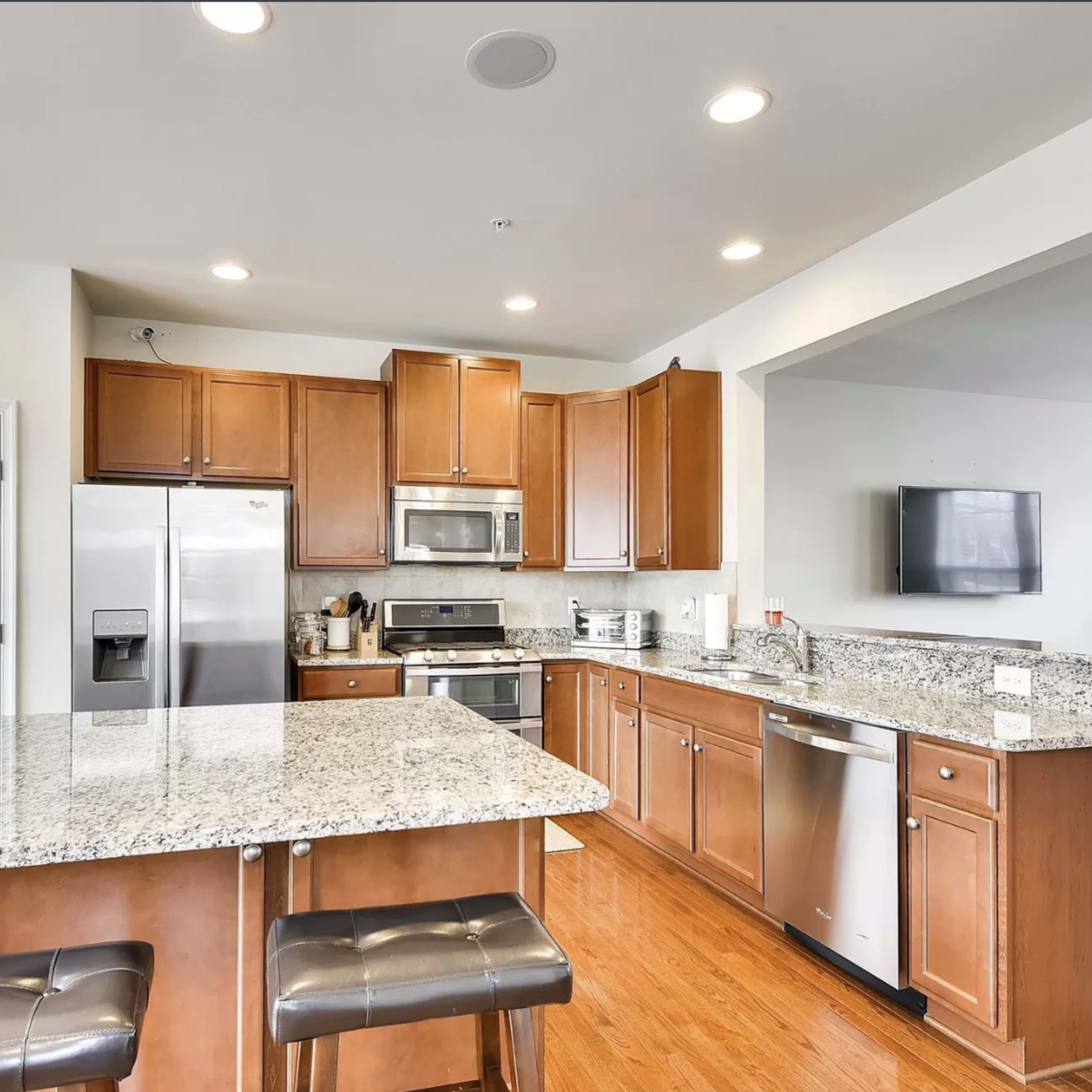 A modern kitchen featuring wooden cabinets, a granite countertop island, stainless steel appliances, and hardwood flooring.