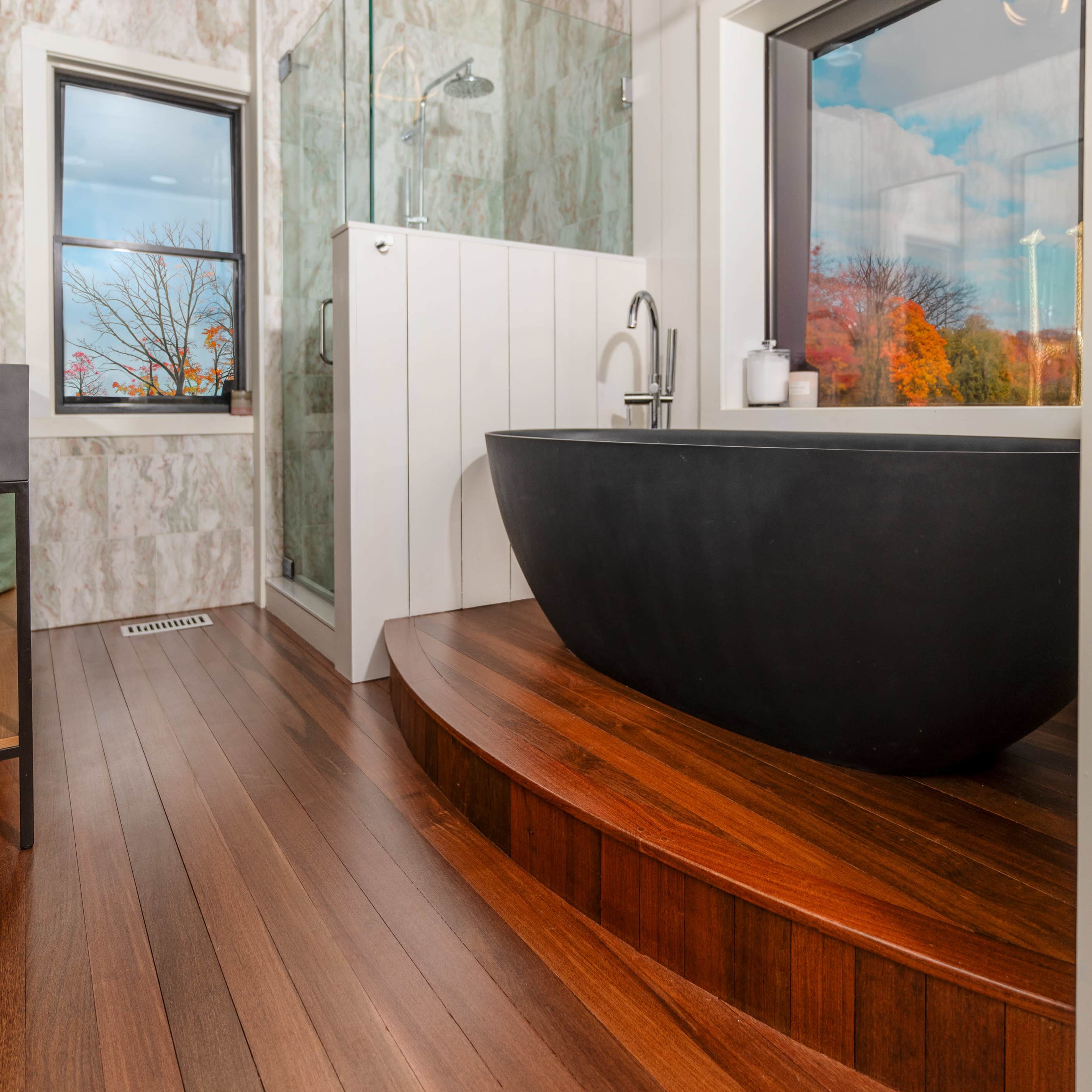 The bathroom features a black freestanding bathtub on a curved wooden platform, with a glass shower enclosure and large windows showcasing an outdoor view.