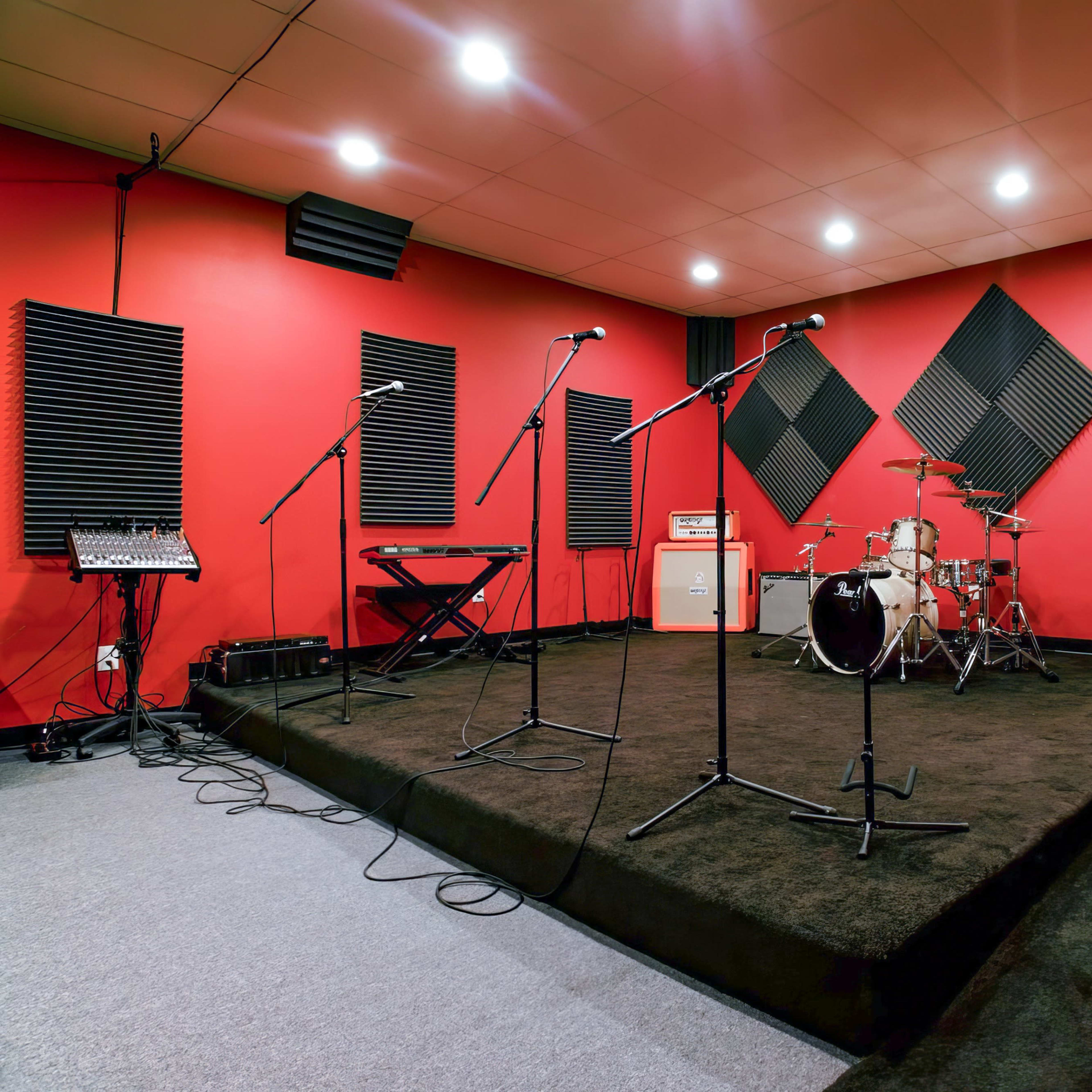 A music rehearsal studio with red walls, black soundproofing panels, a drum set, keyboard, and amplifiers arranged on a dark carpeted floor.