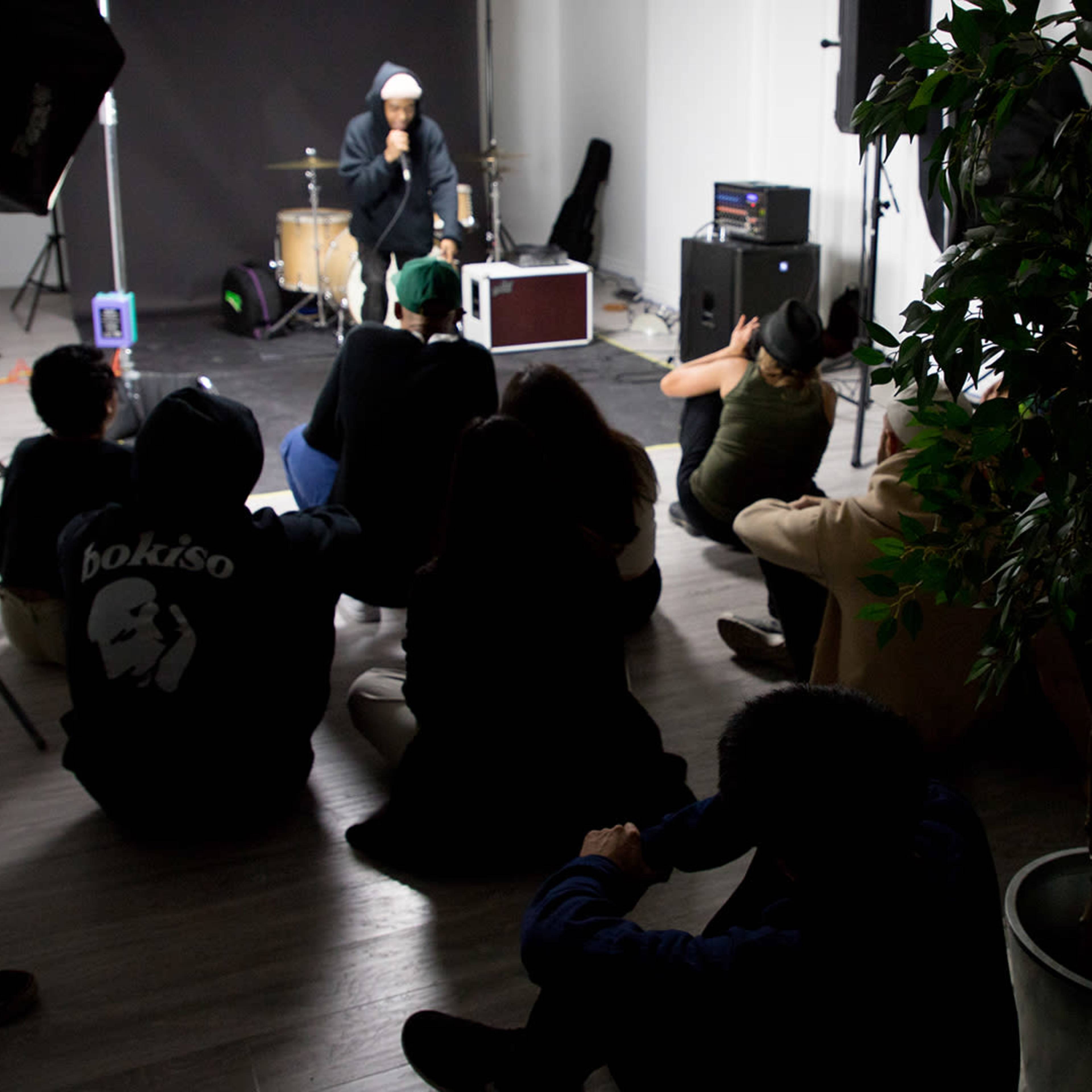 A musician performs in a studio while an audience sits on the floor, listening attentively.