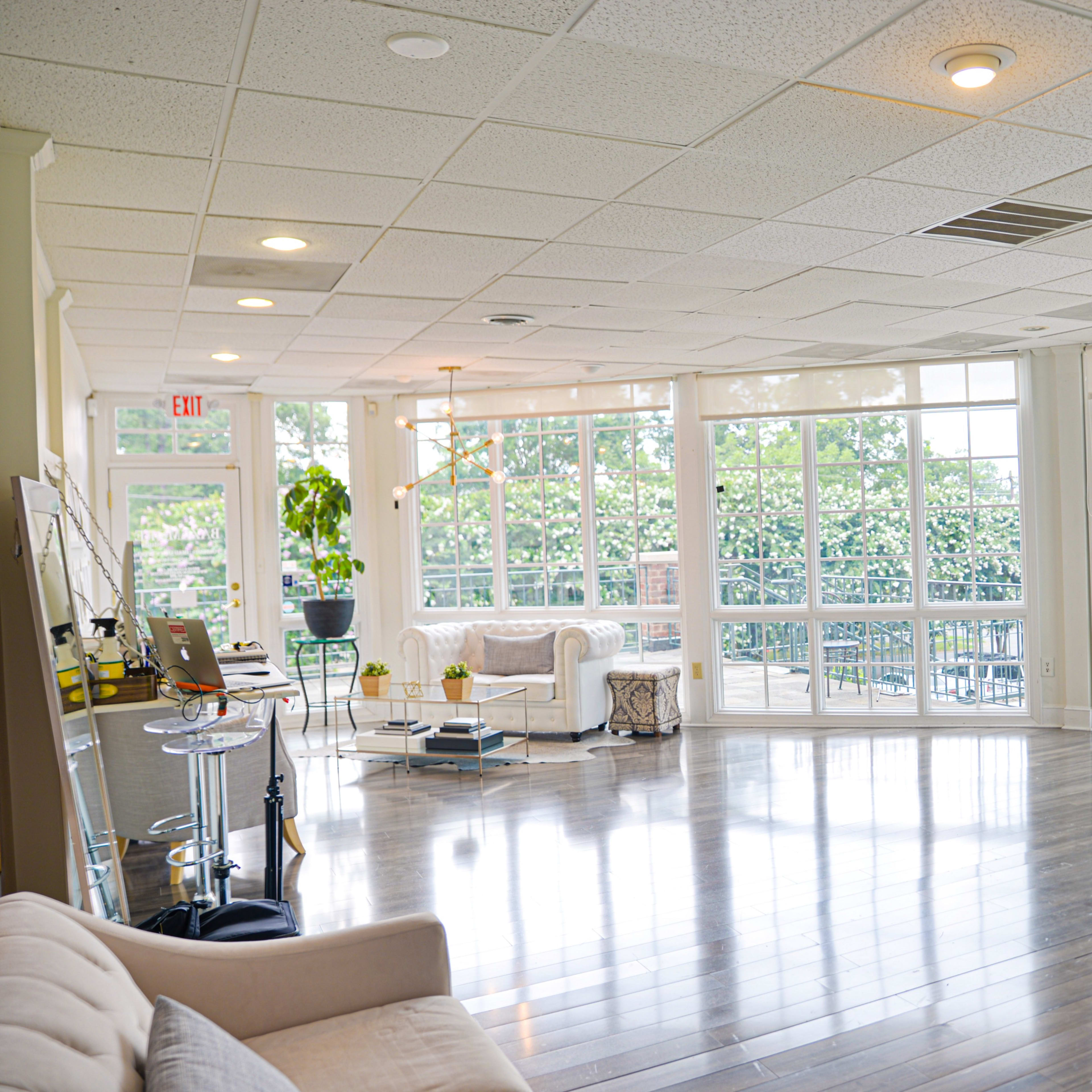 A spacious room with large windows allowing natural light, hardwood flooring, and minimal furnishings including a couch and a table with a laptop.