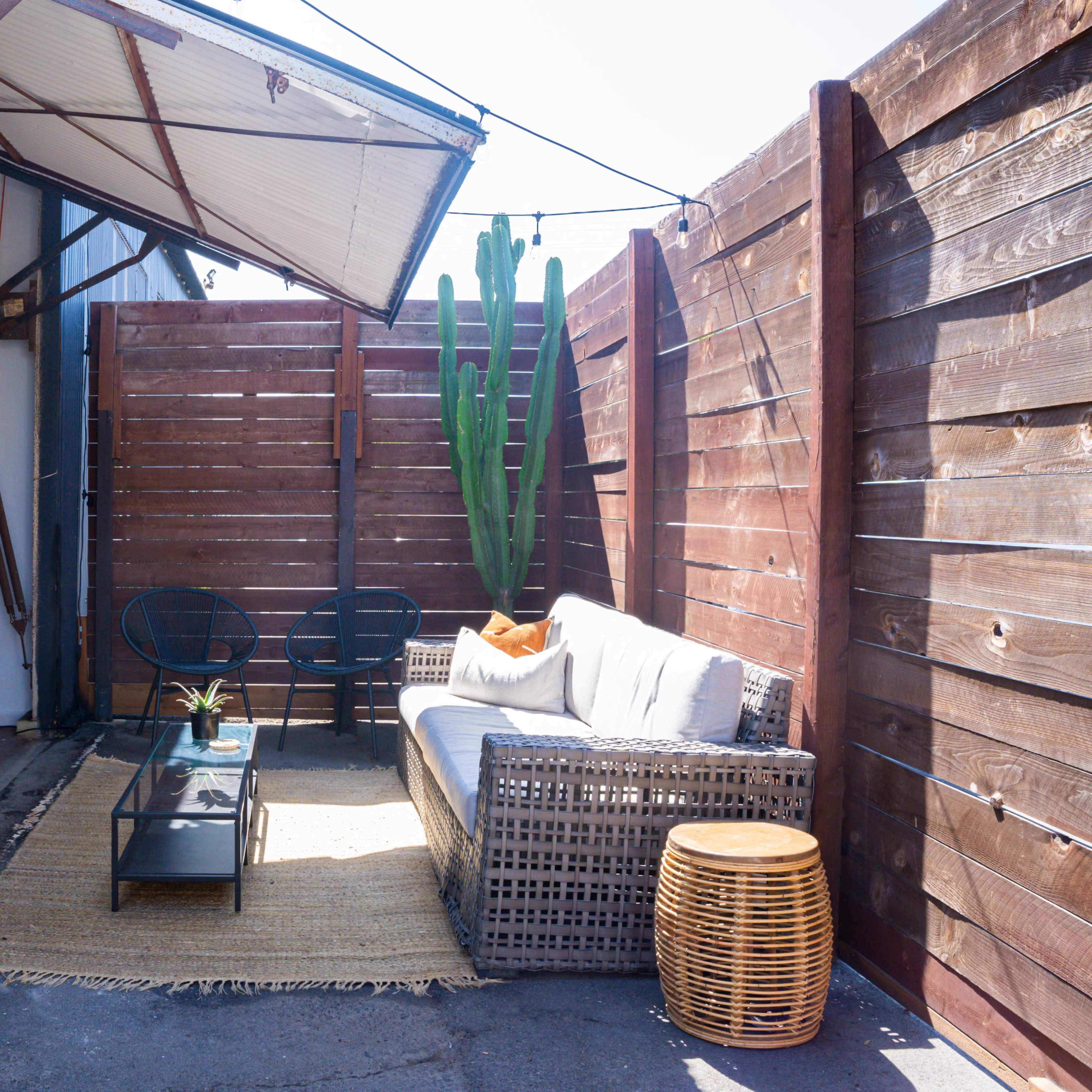 A small outdoor seating area with a wicker sofa, a coffee table, and a potted cactus, enclosed by wooden fences and a metal building.