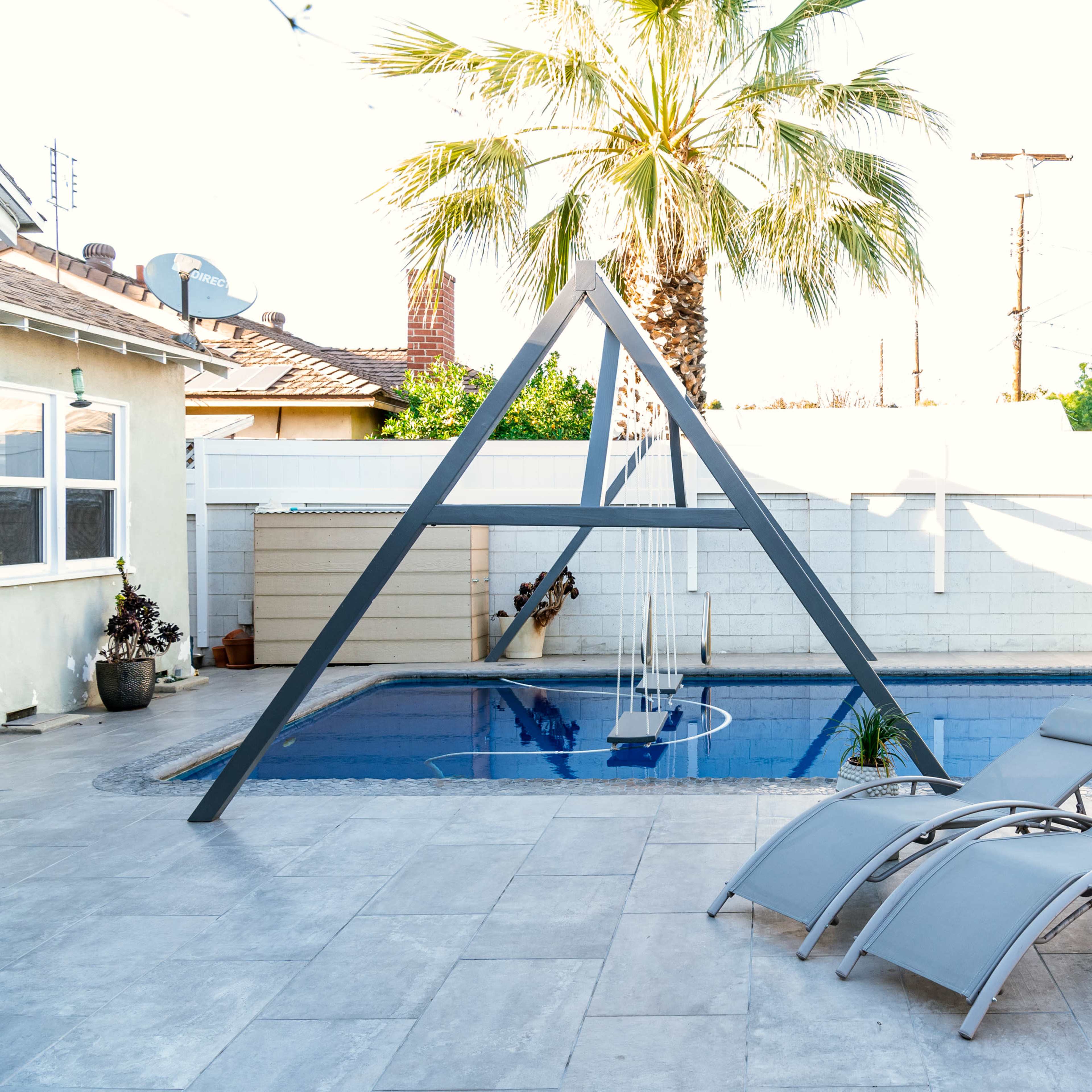 A backyard pool area featuring an A-frame structure, two lounge chairs, and a palm tree.