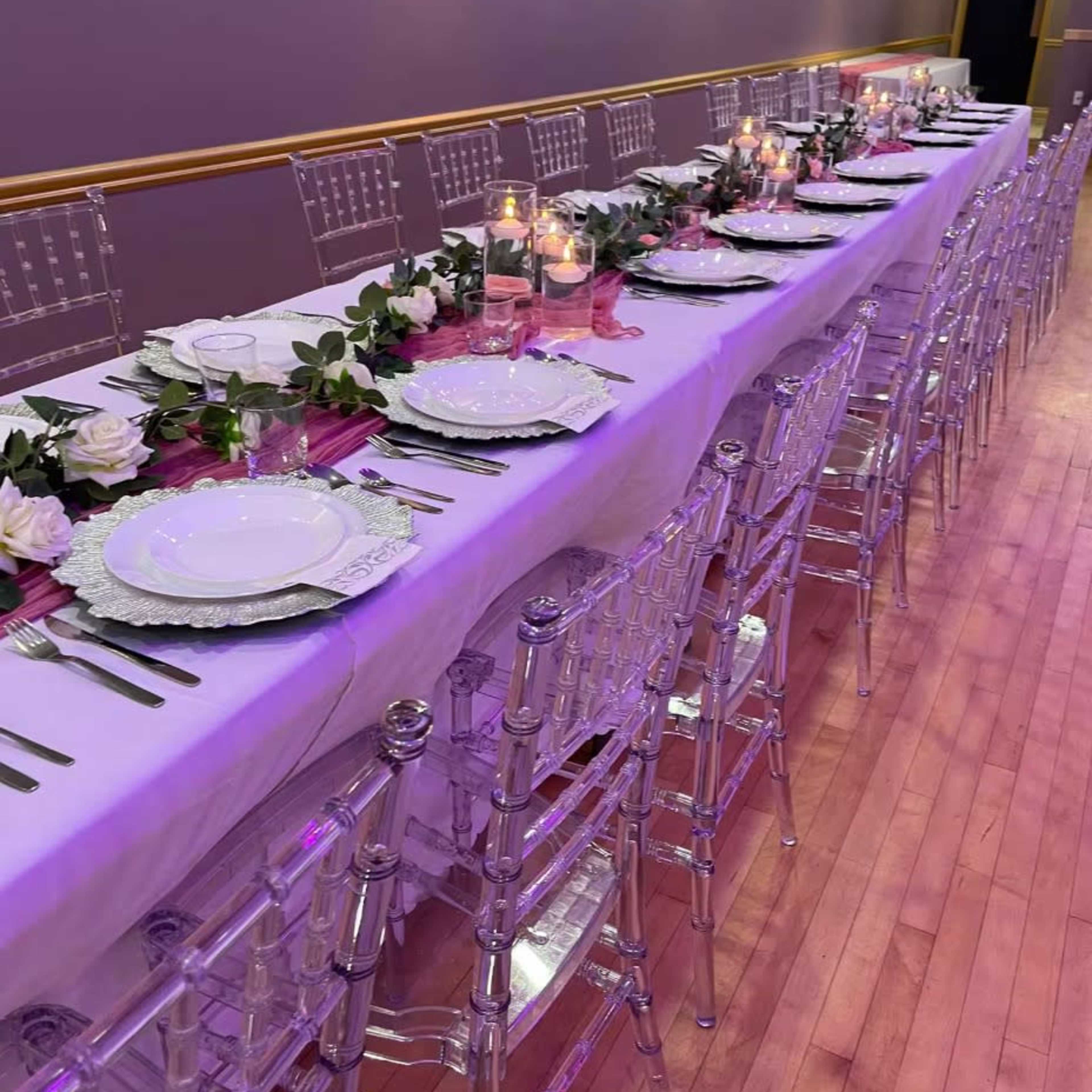 A long banquet table is set with white plates, silverware, and candles, decorated with greenery and white roses, under soft lighting.