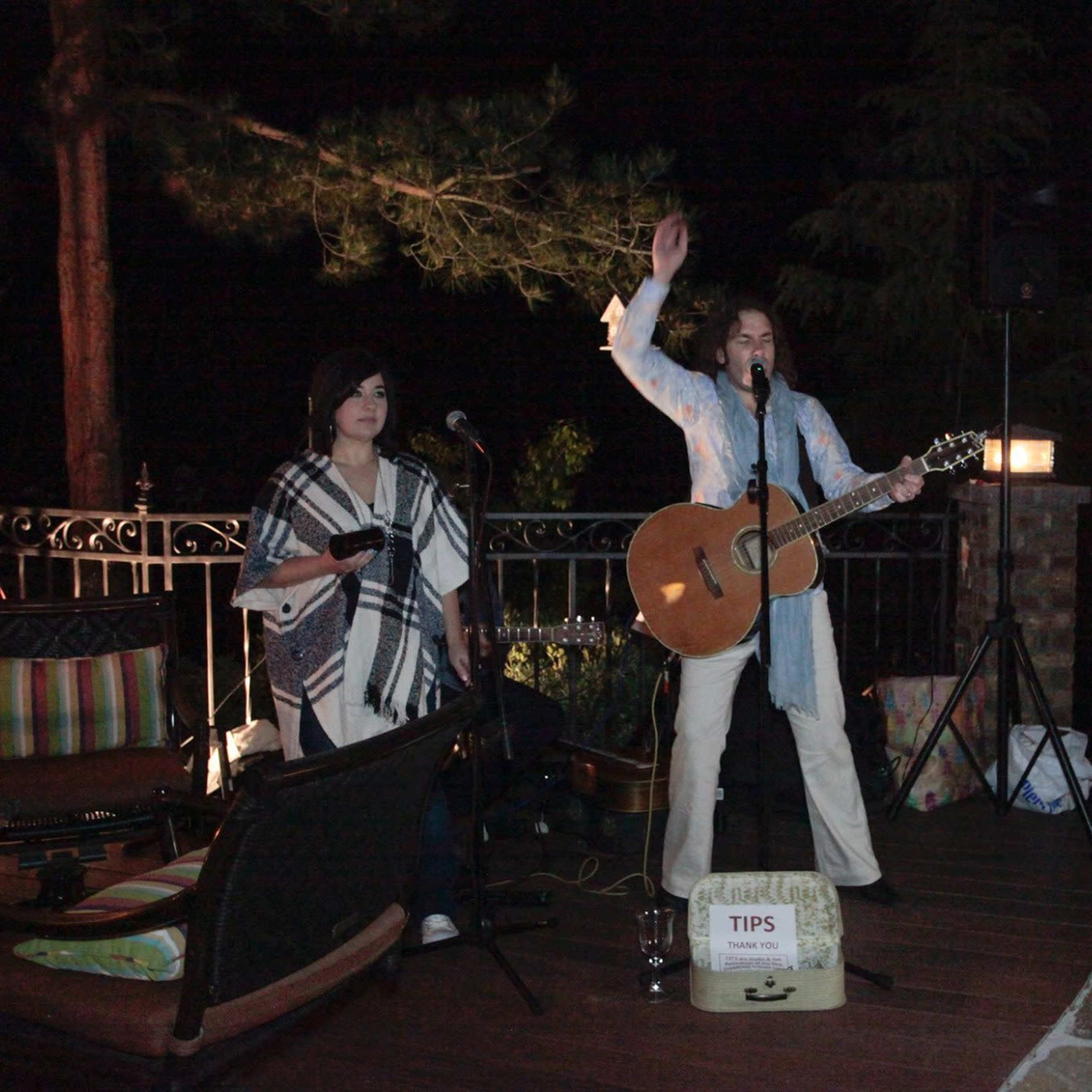 Two musicians perform on a deck at night, with one playing guitar and the other standing beside a tips box.