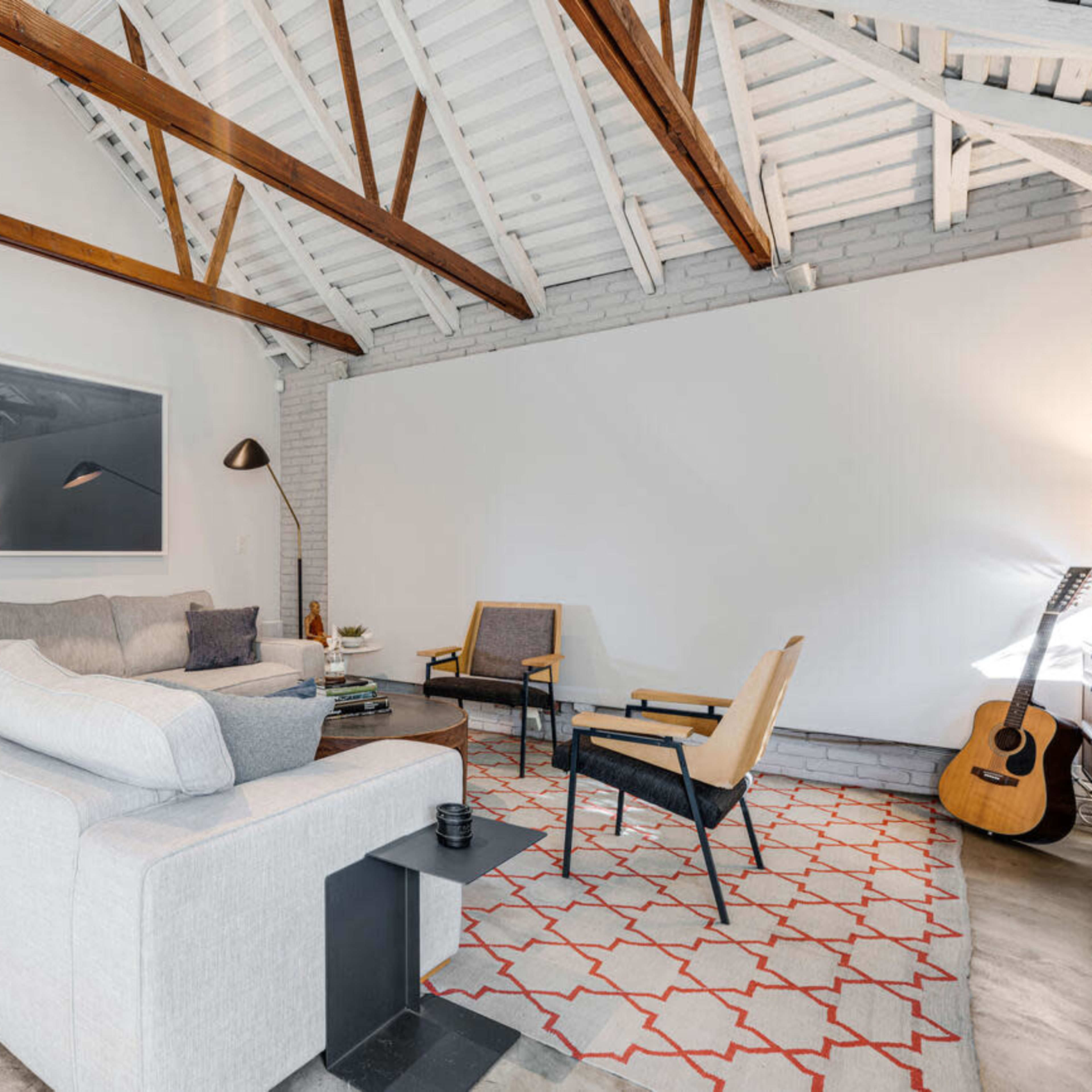 A modern living room with a large sectional sofa, a coffee table, and an acoustic guitar in a corner, featuring exposed wooden beams and a patterned area rug.