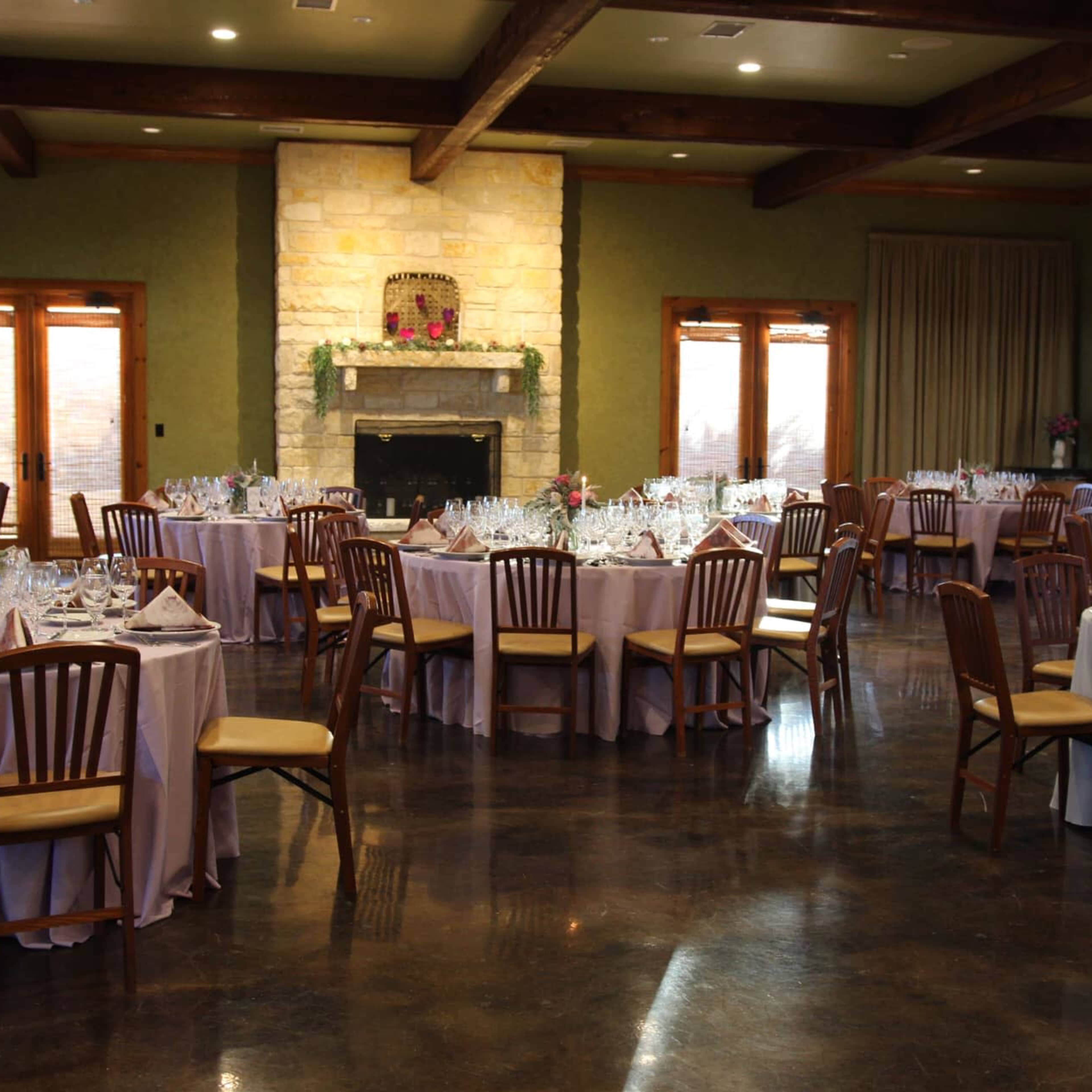 A spacious event room features several round tables dressed in pastel tablecloths, with an elegant fireplace and large windows in the background.