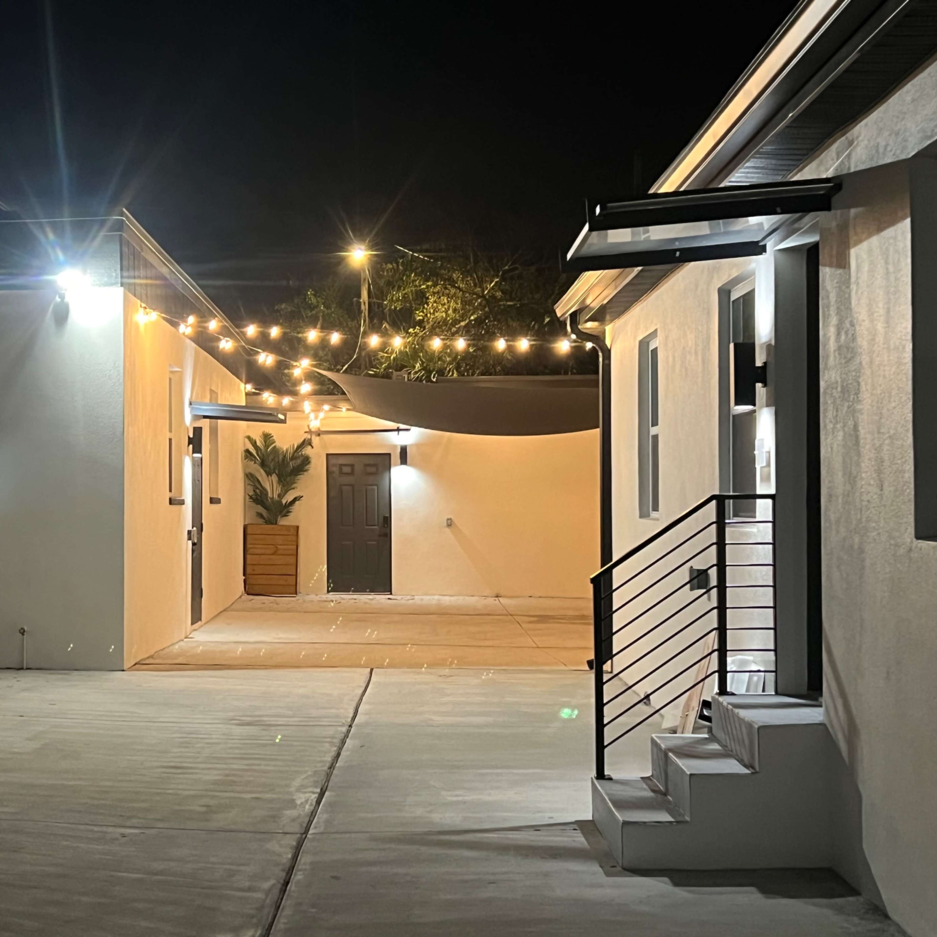 A well-lit courtyard connects two buildings with string lights overhead and potted plants beside a door.