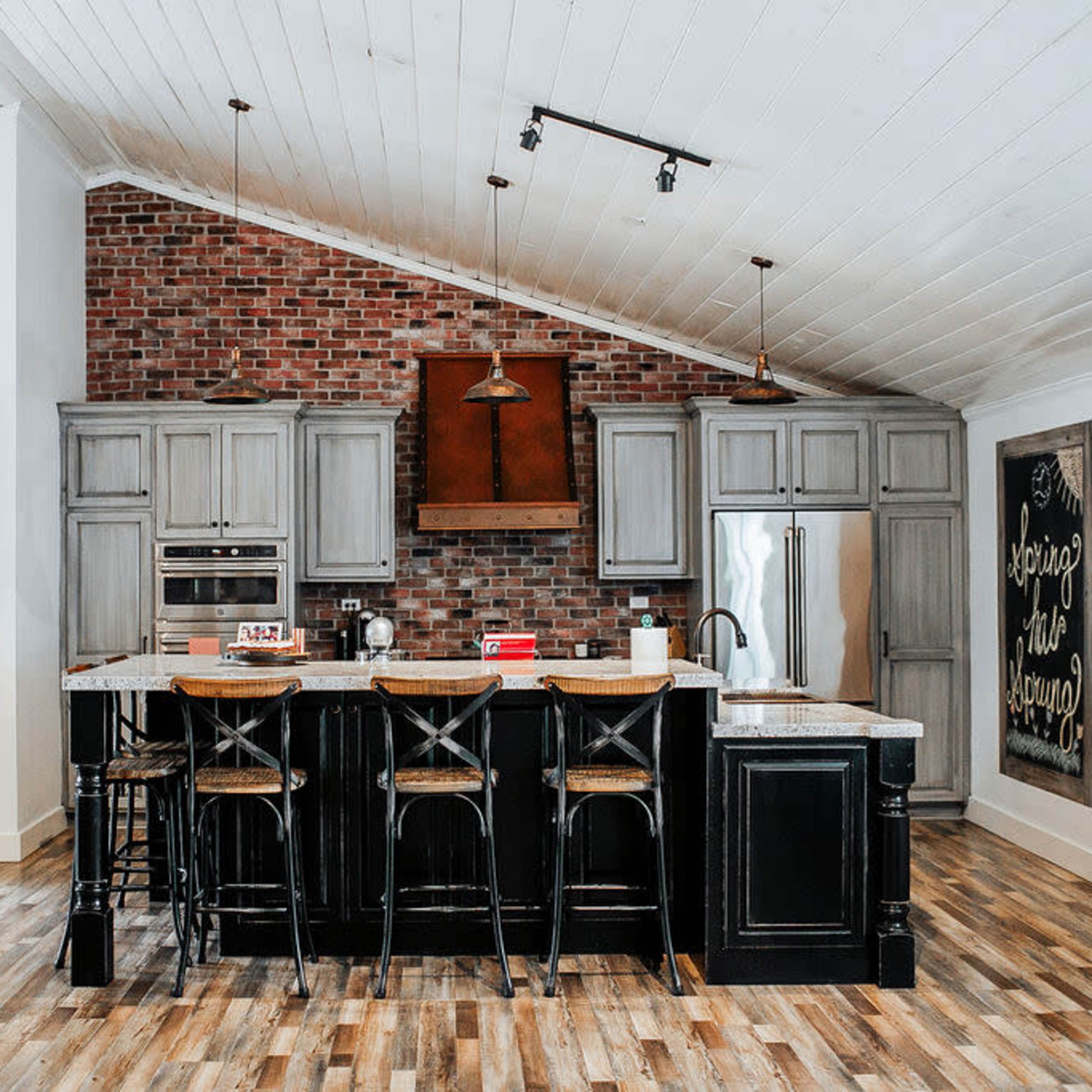 A modern kitchen featuring gray cabinets, a brick accent wall, and a large island with bar stools.