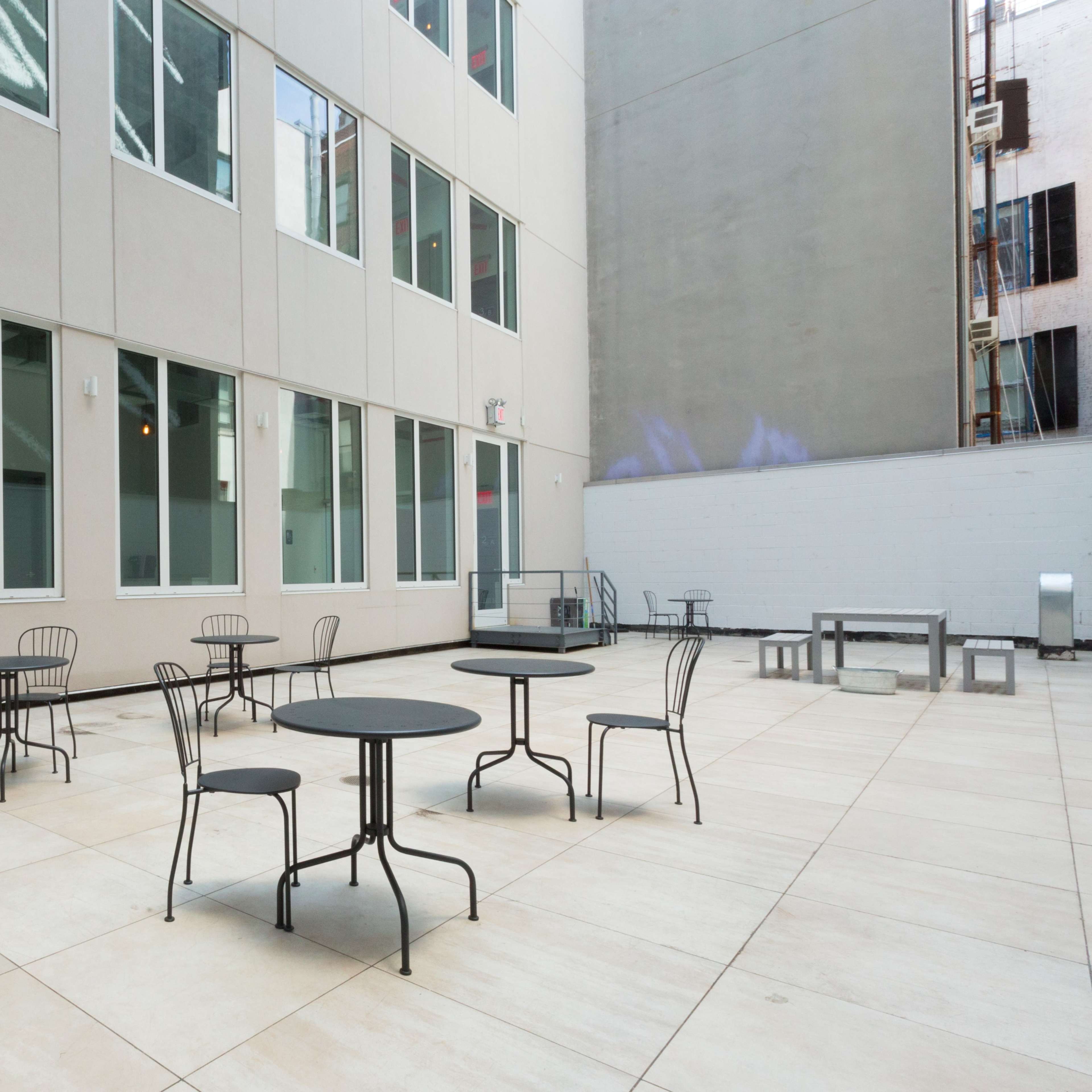 An outdoor courtyard with several small tables and chairs situated on a tiled surface, surrounded by tall building walls.