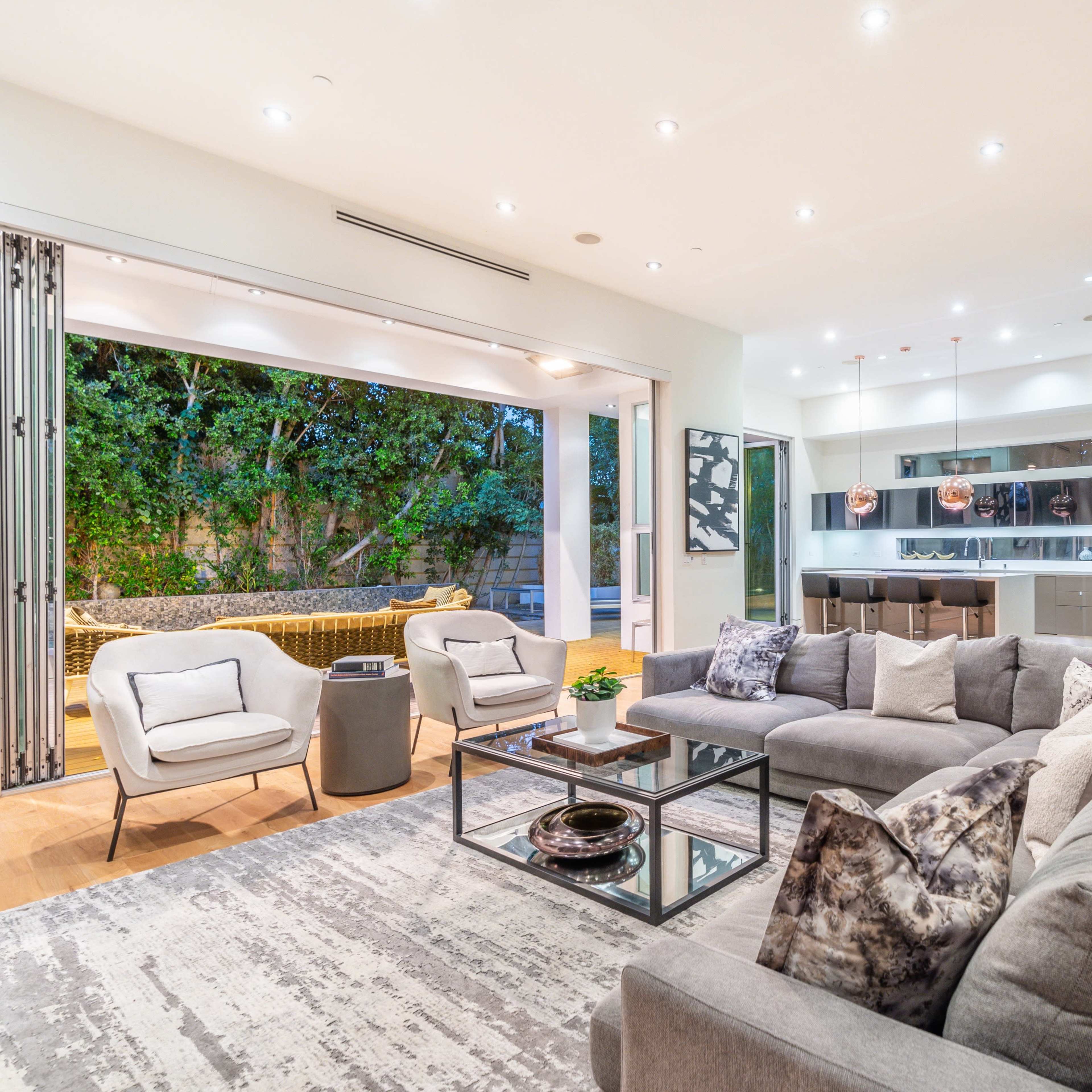A modern living room with a large sectional sofa, a glass coffee table, and an open view into an outdoor area framed by sliding glass doors.