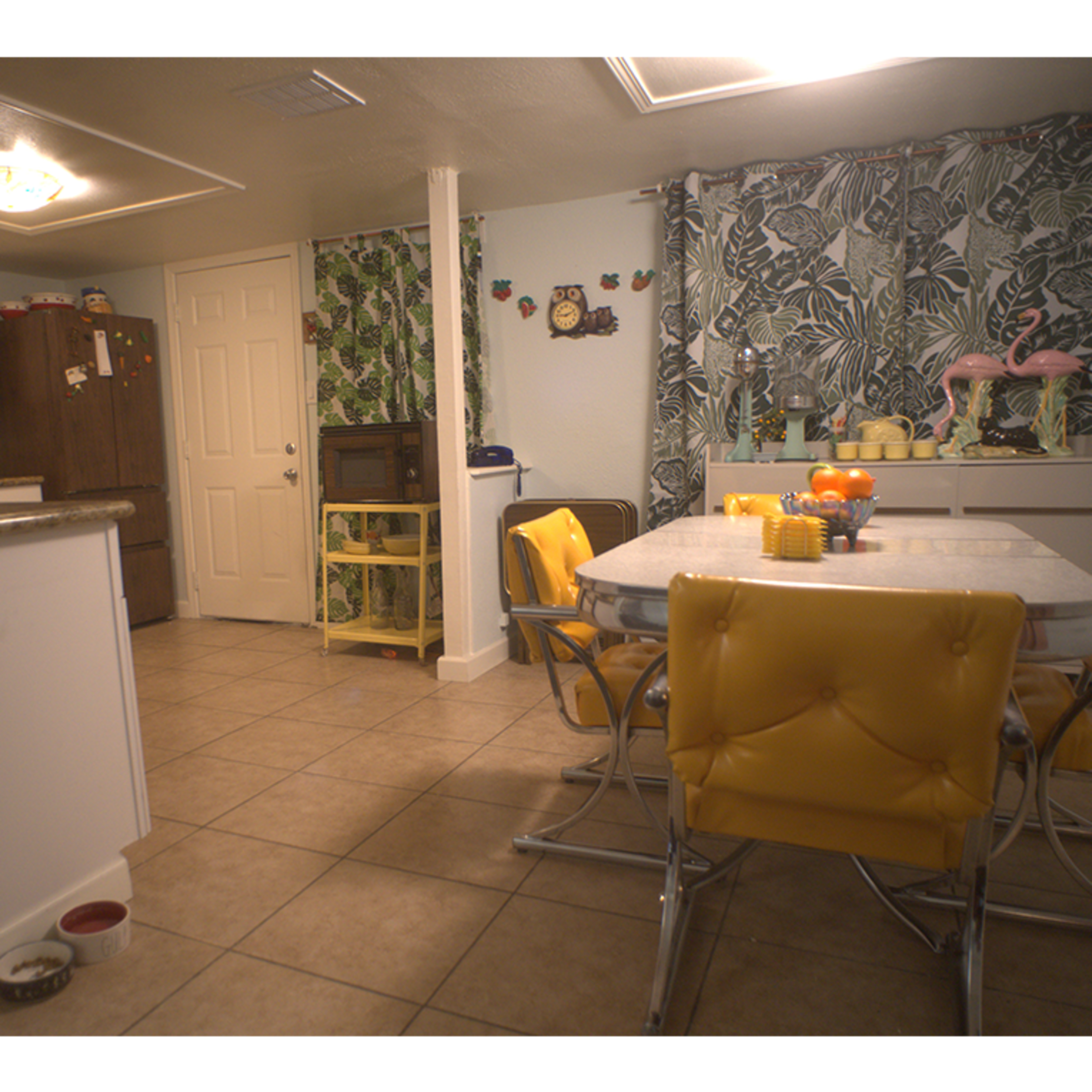 A kitchen area with a table surrounded by yellow chairs, tropical-patterned curtains, and various items on the countertops.
