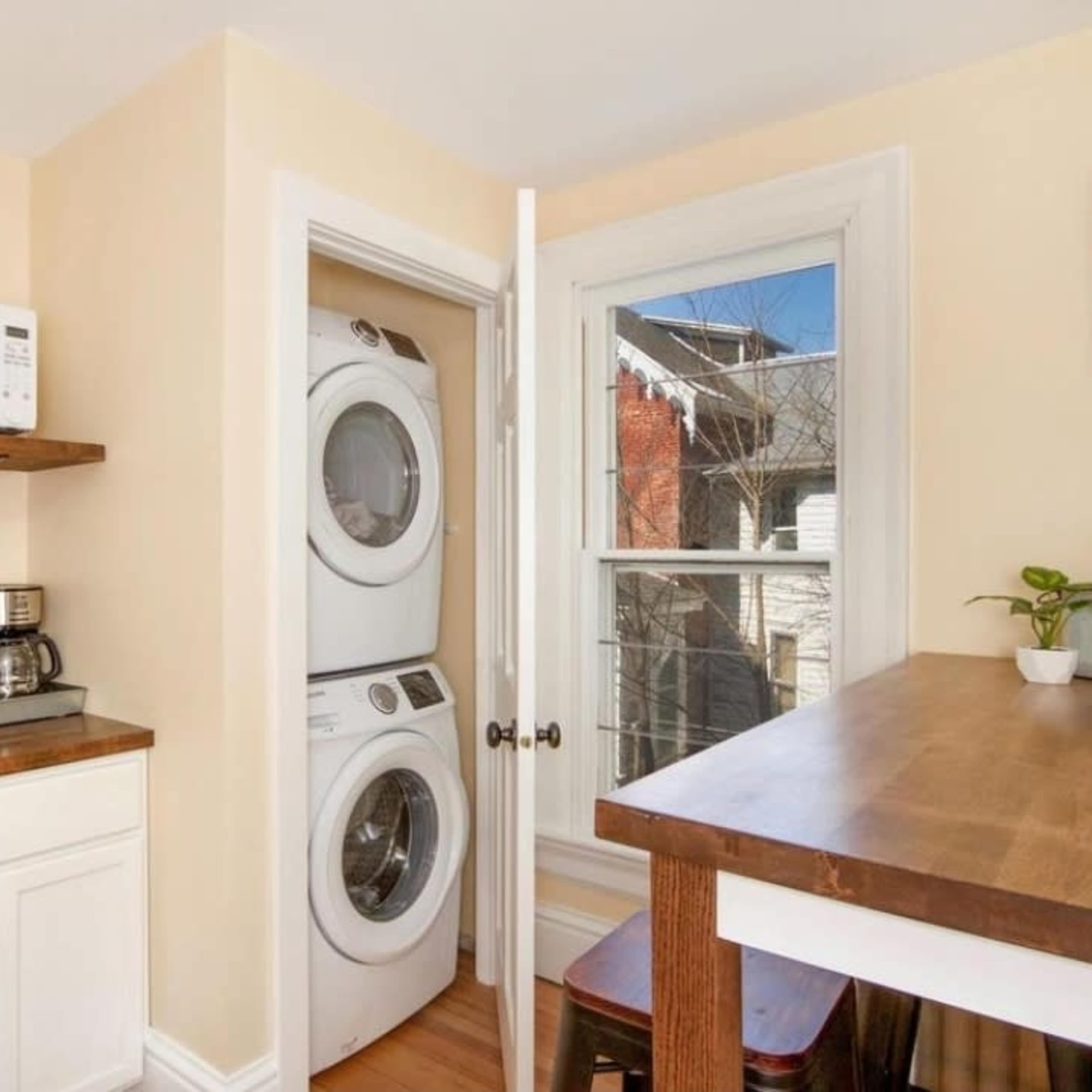A small laundry area with a stacked washer and dryer next to a kitchen counter with a coffee maker and a microwave, adjacent to a window.