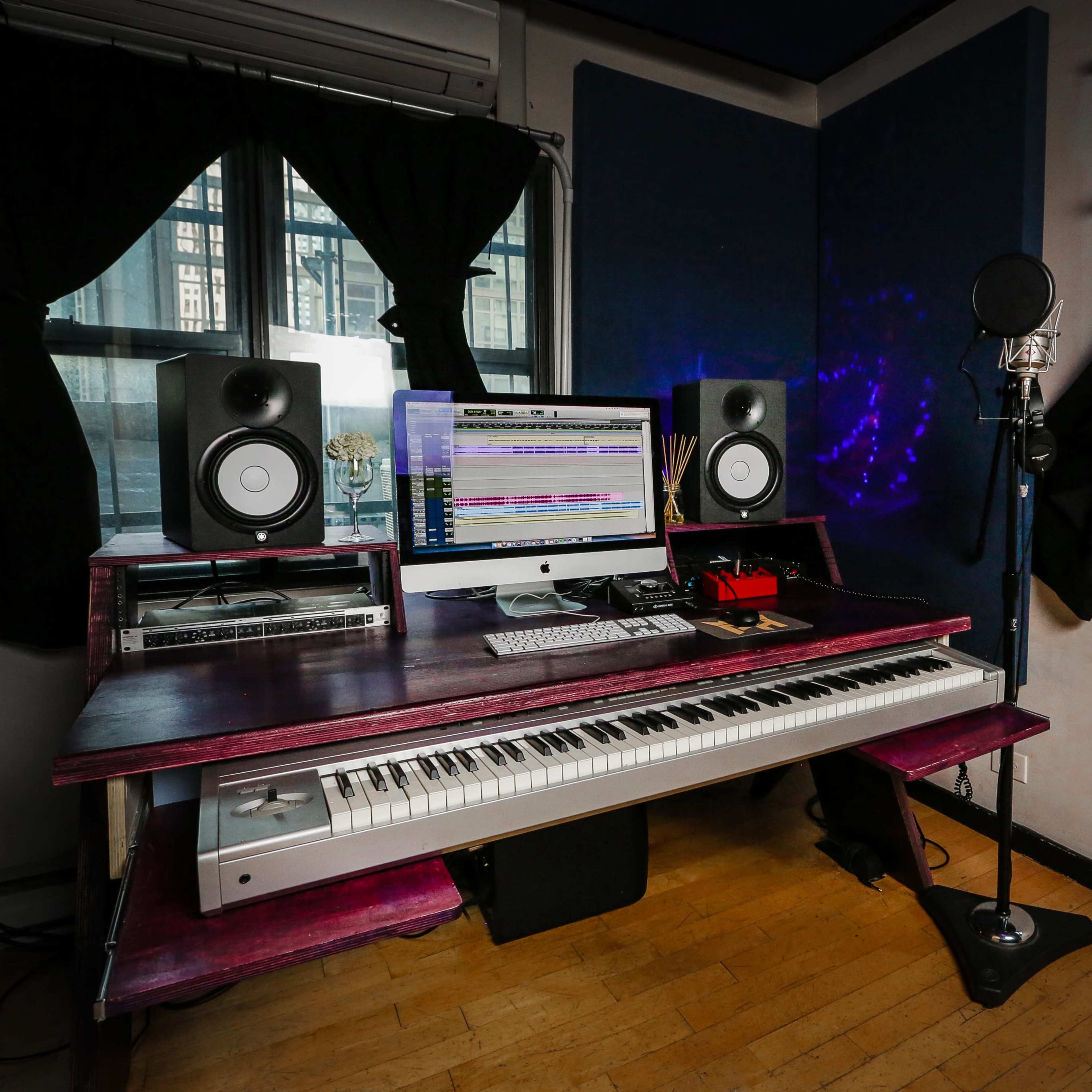 A compact music studio featuring a computer with audio software, studio monitors, a microphone on a stand, and a keyboard, set against a window view and soundproofing panels.