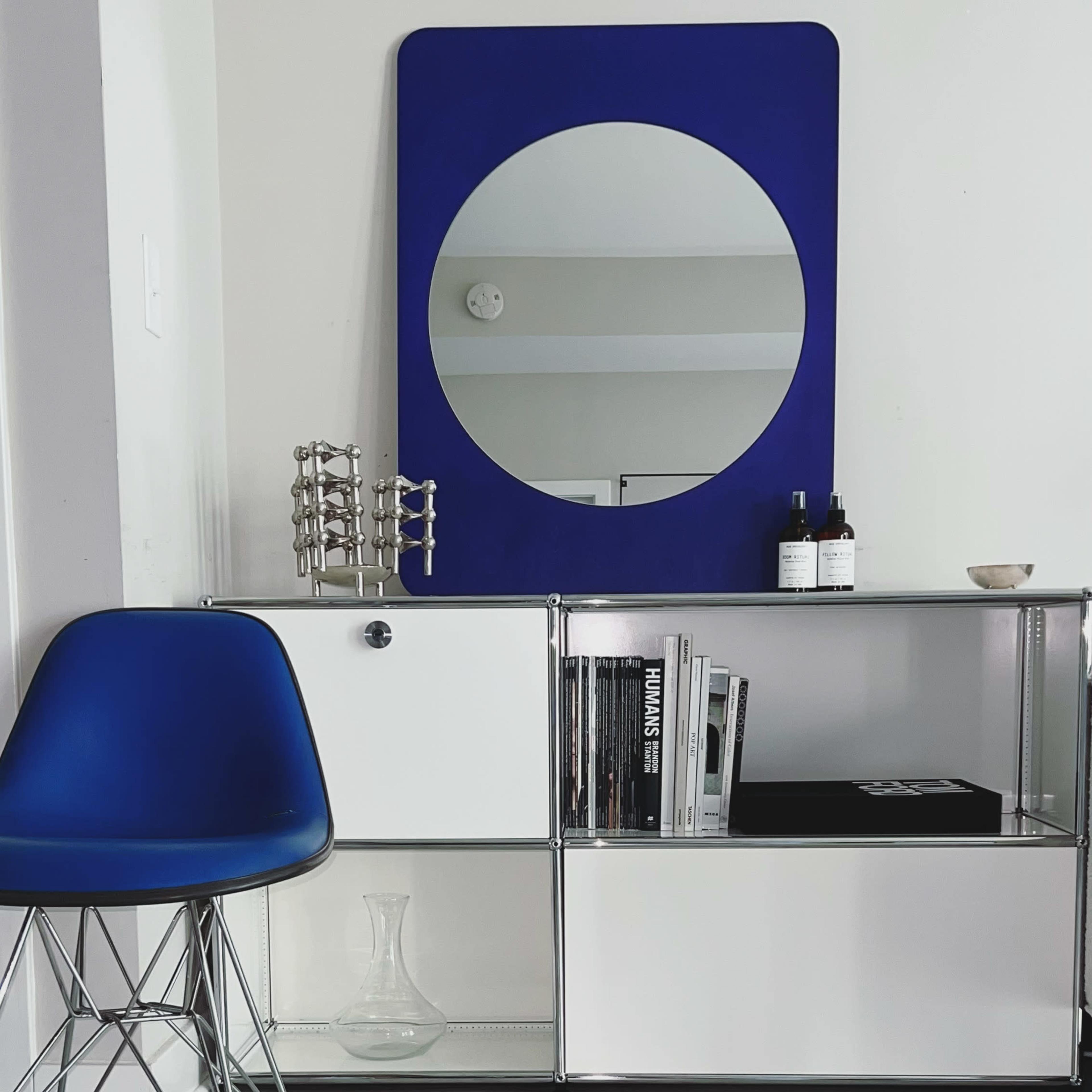 A modern interior scene featuring a blue chair next to a white and chrome shelving unit with a circular mirror above it.