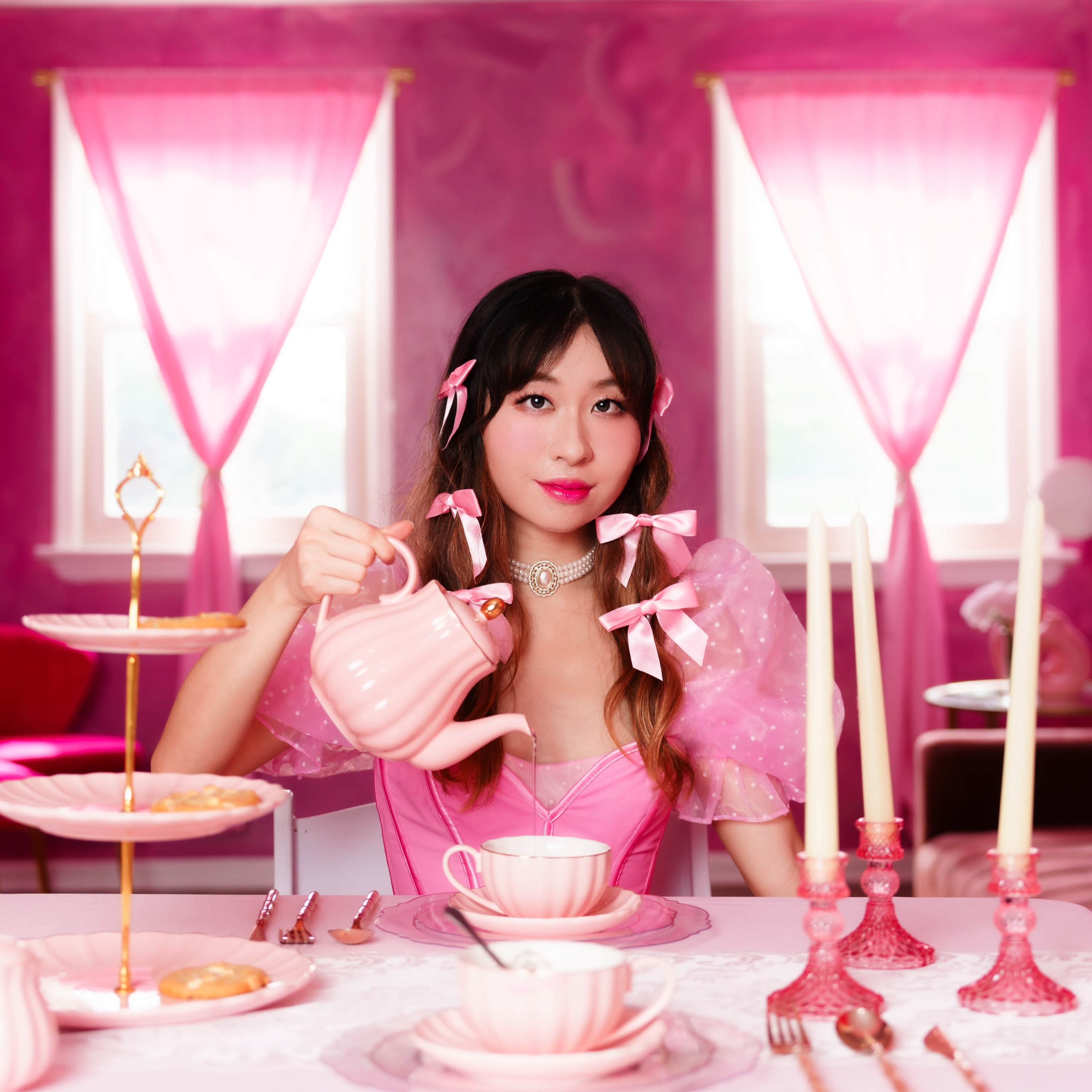 A woman in a pink outfit pours tea from a matching teapot at a table set with pink dishware and decorative candles in a pink-themed room.