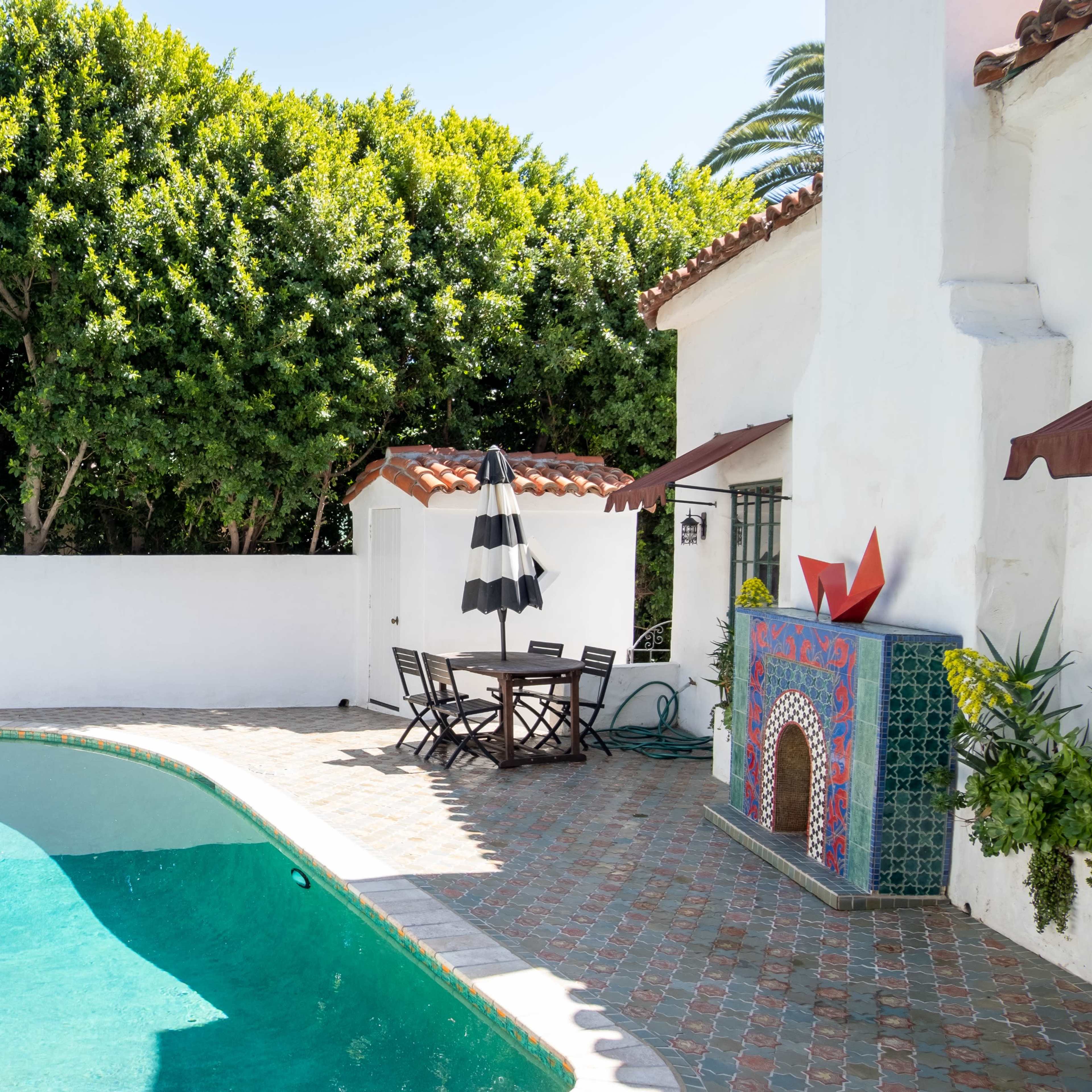 A pool area surrounded by greenery, with a tiled BBQ and outdoor dining table under an umbrella beside the pool.