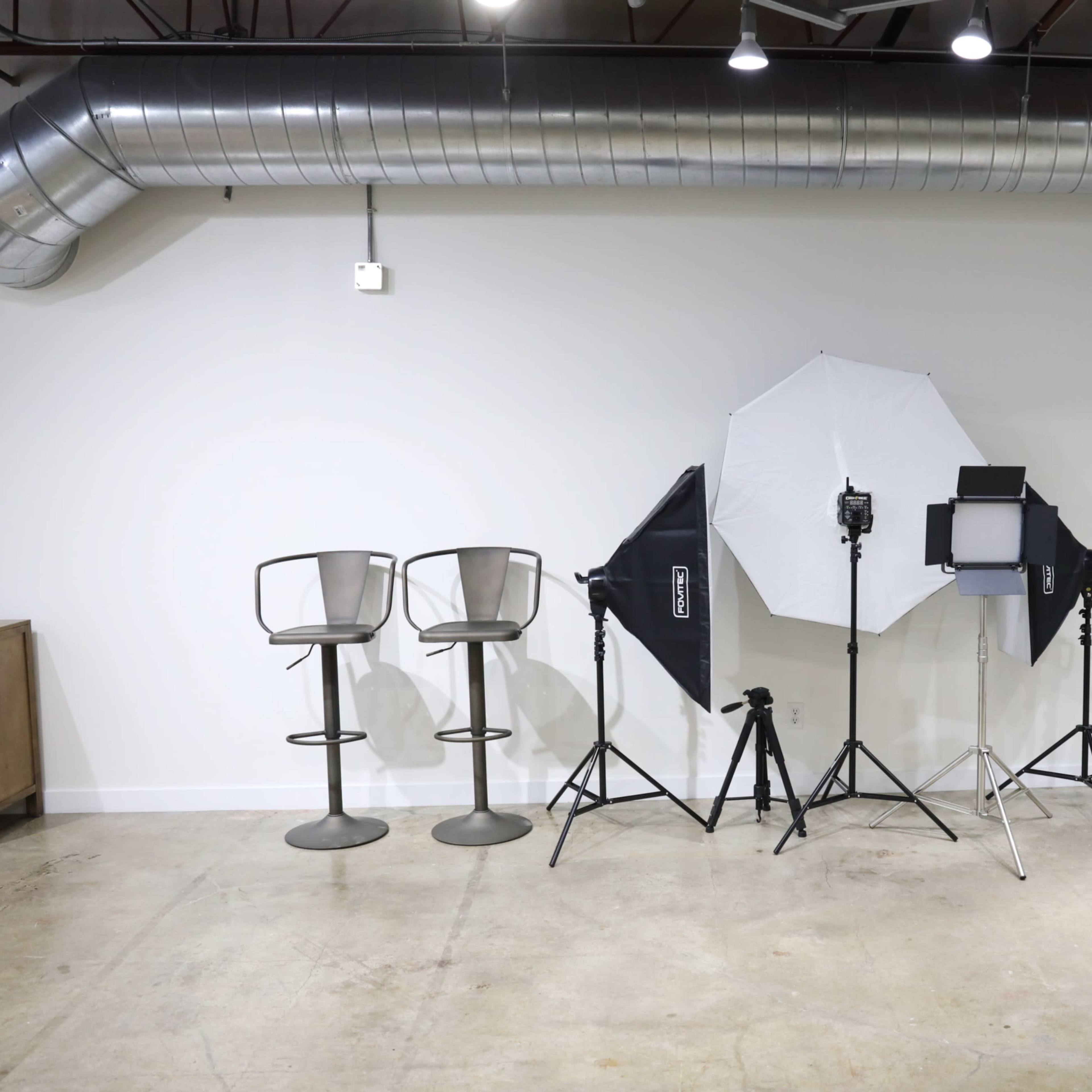 A blank interior space with two bar stools and three studio lighting equipment setups against a white wall.
