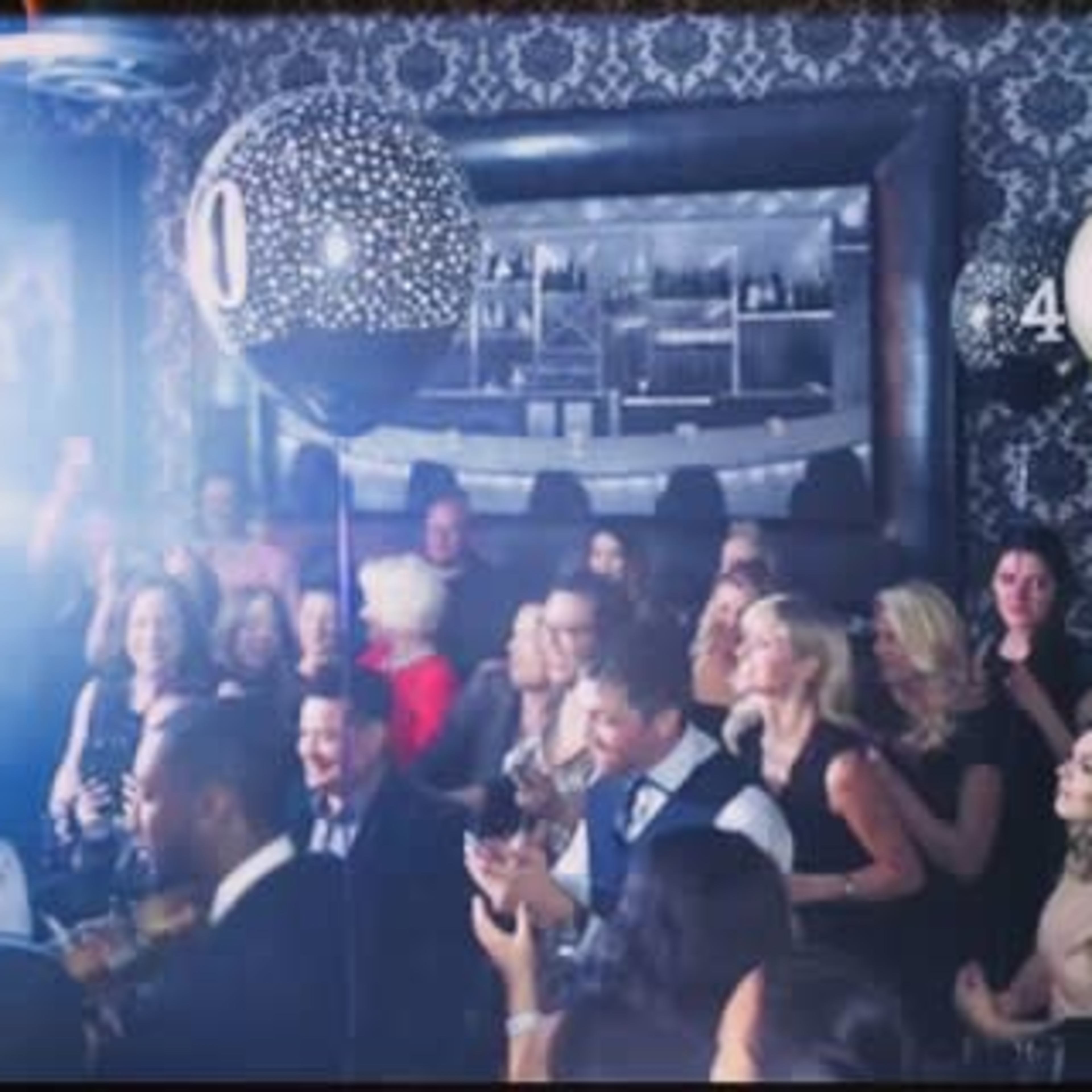 A crowd of people celebrates in a dimly lit venue decorated with balloons.