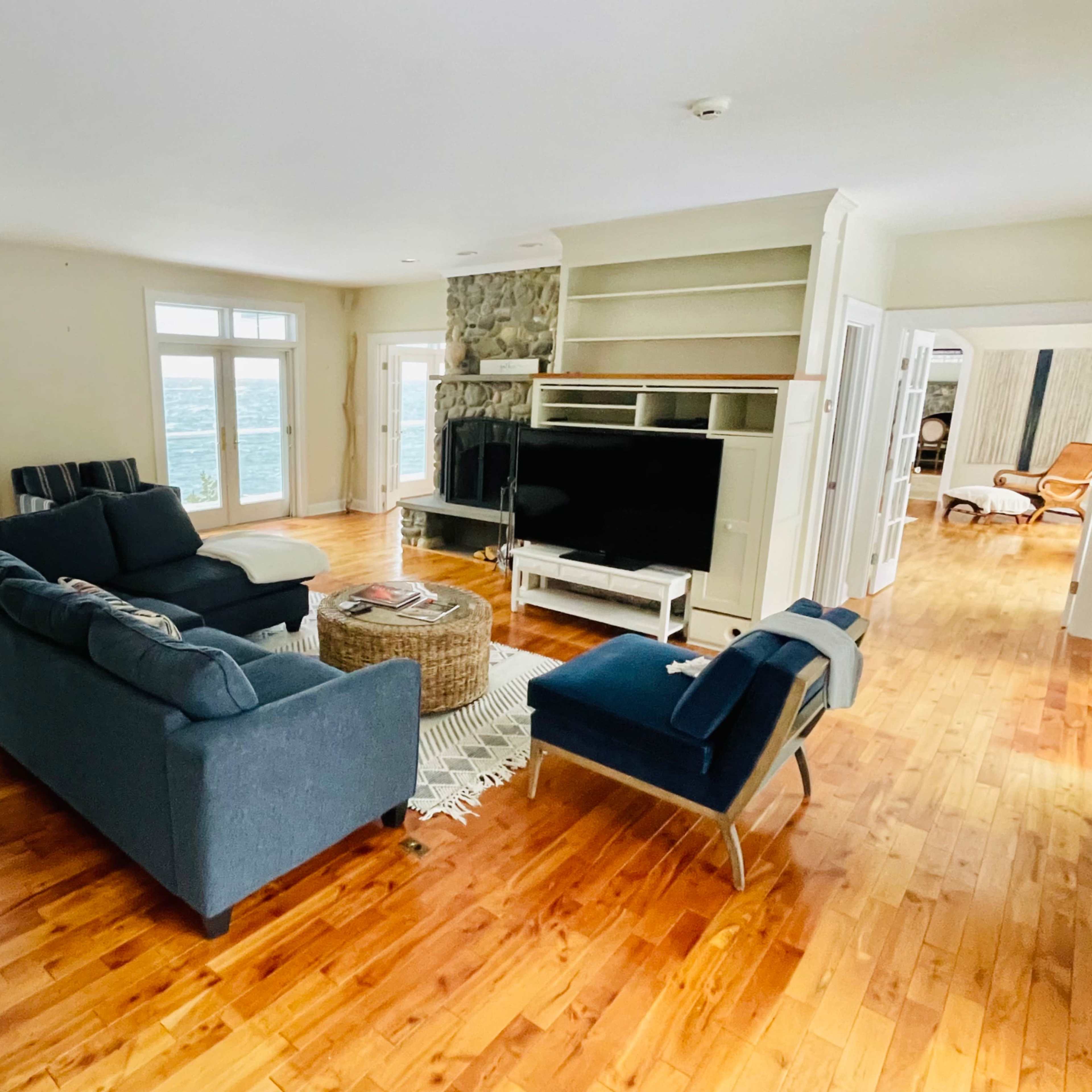 The living room features a blue sectional sofa, a coffee table, and a stone fireplace, with large windows overlooking a body of water.