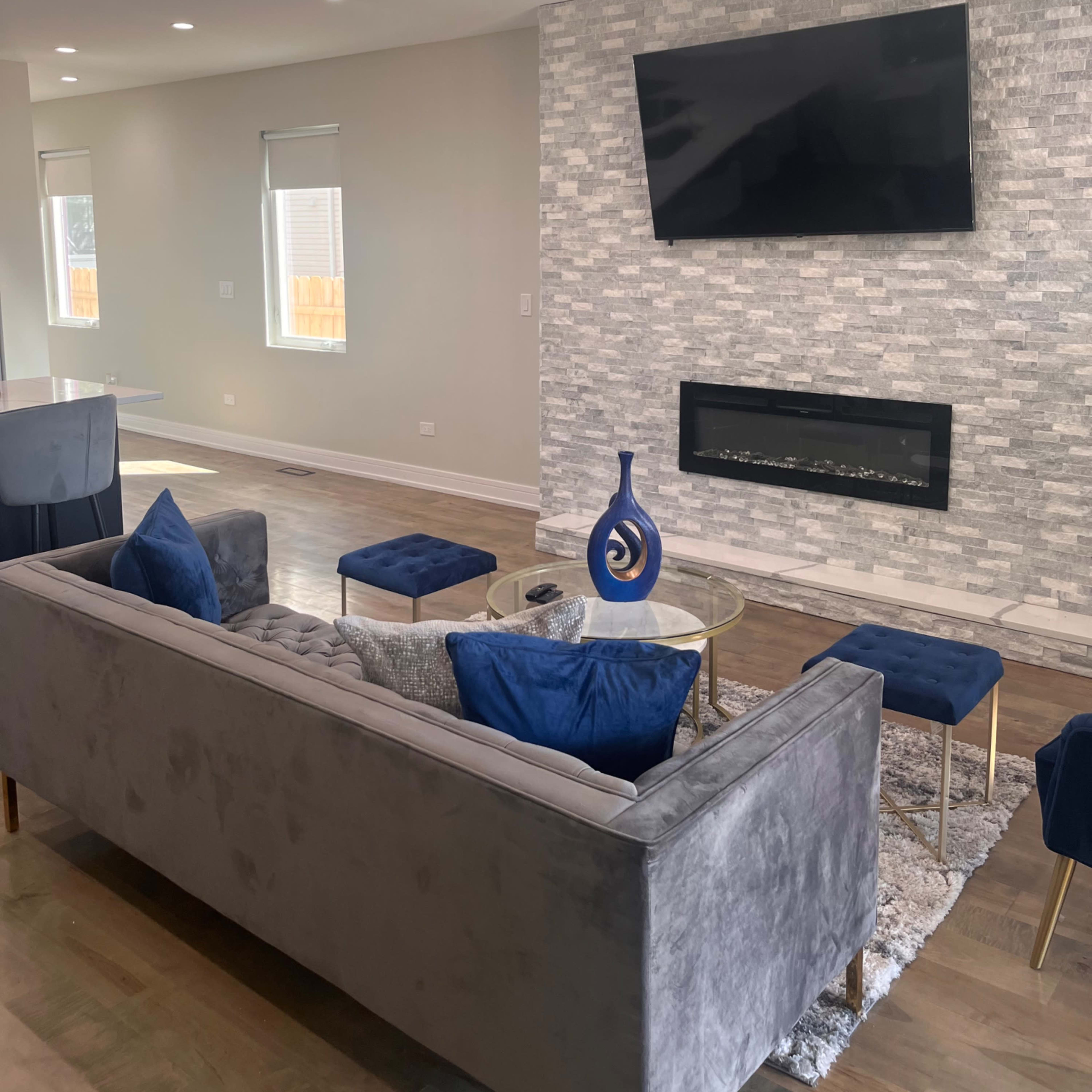The living room features a gray sofa with blue accent pillows, a glass coffee table, and a wall-mounted television above a modern fireplace, set against a textured stone background.