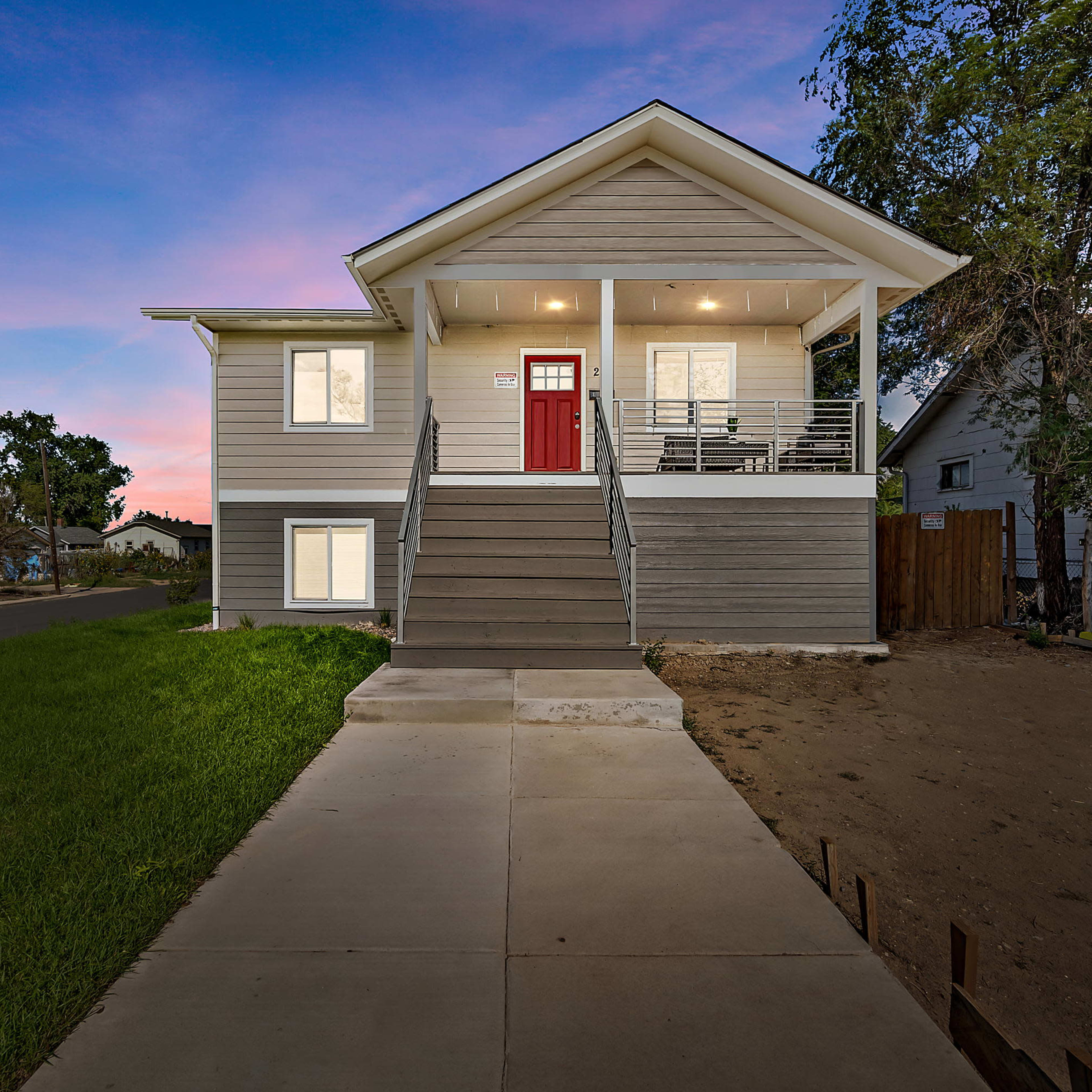 A modern gray house with a red front door and a staircase, situated on a grassy yard beside a concrete walkway.