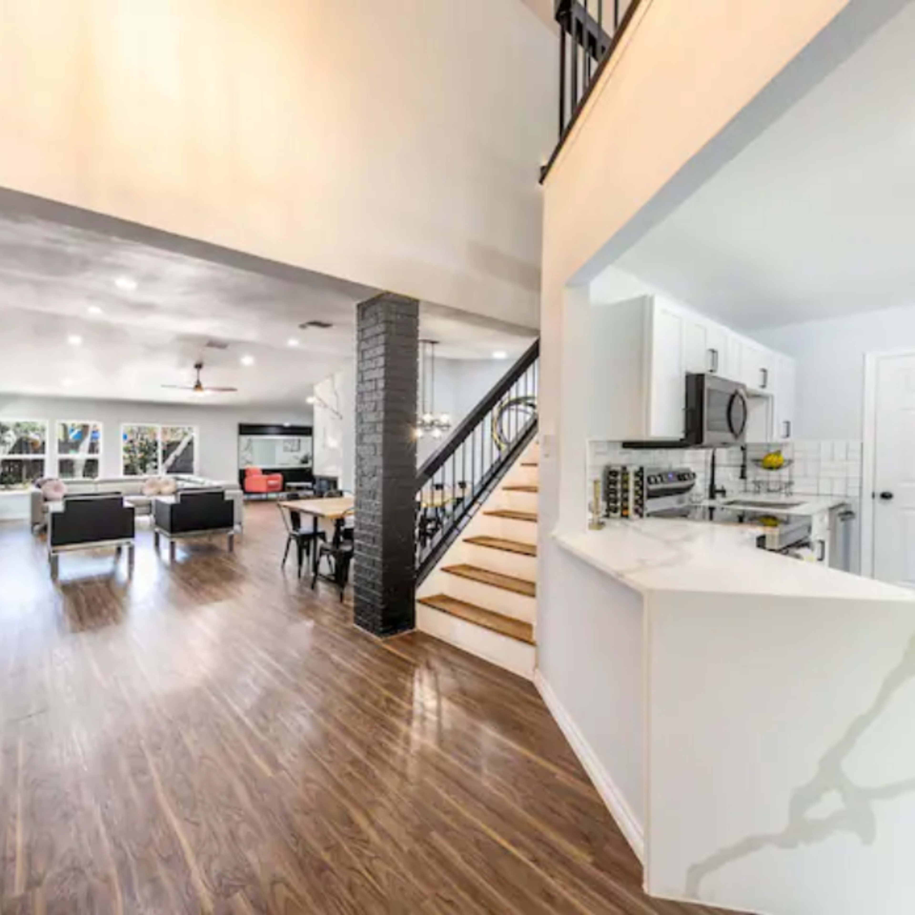 A modern interior space with an open layout, featuring a staircase on the right, a kitchen area, and a living area with seating and a dining table.