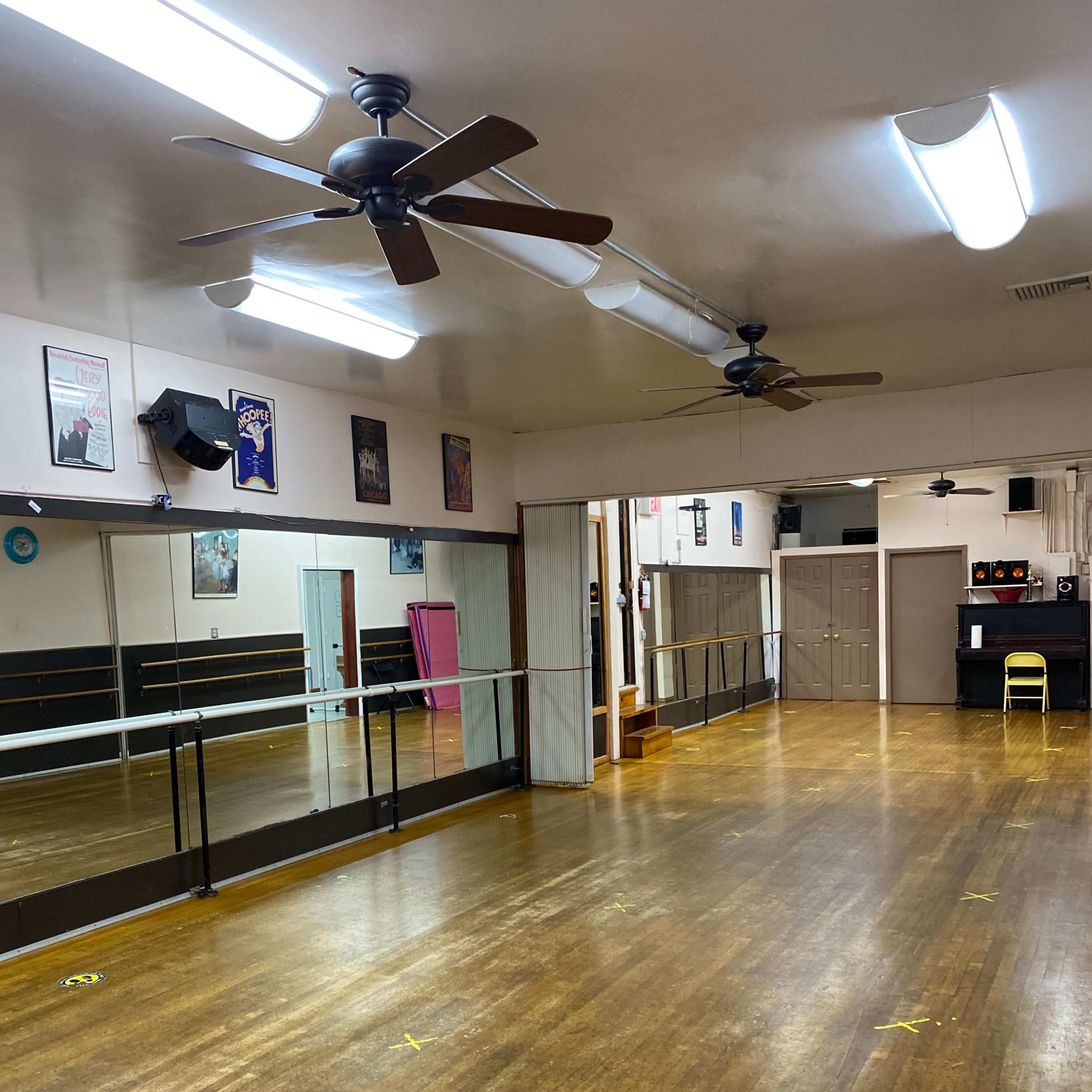 A spacious dance studio with wooden flooring, mirrors on one wall, and a few chairs arranged along the sides.