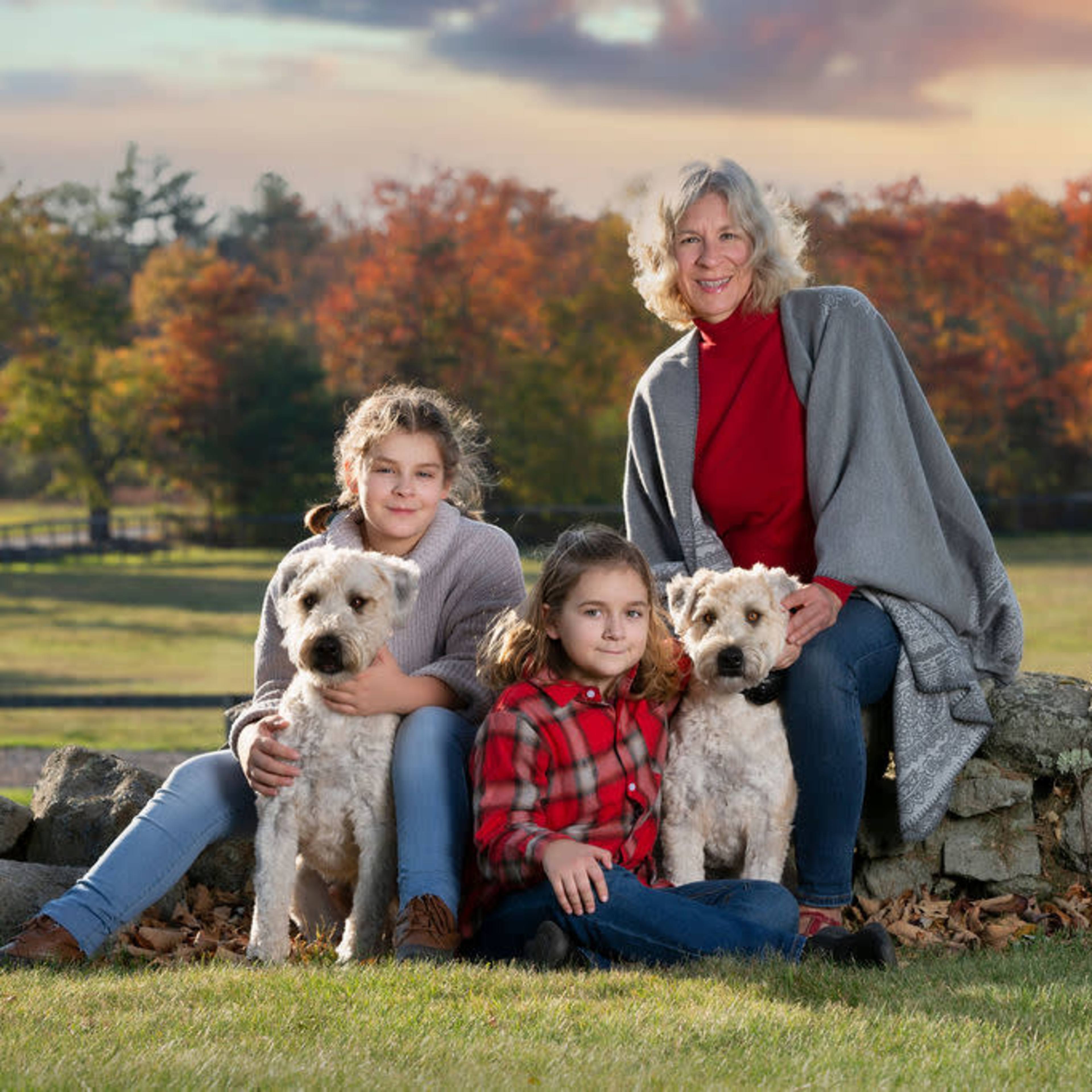 A woman and two girls sit on a stone wall with two dogs, surrounded by a picturesque autumn landscape.