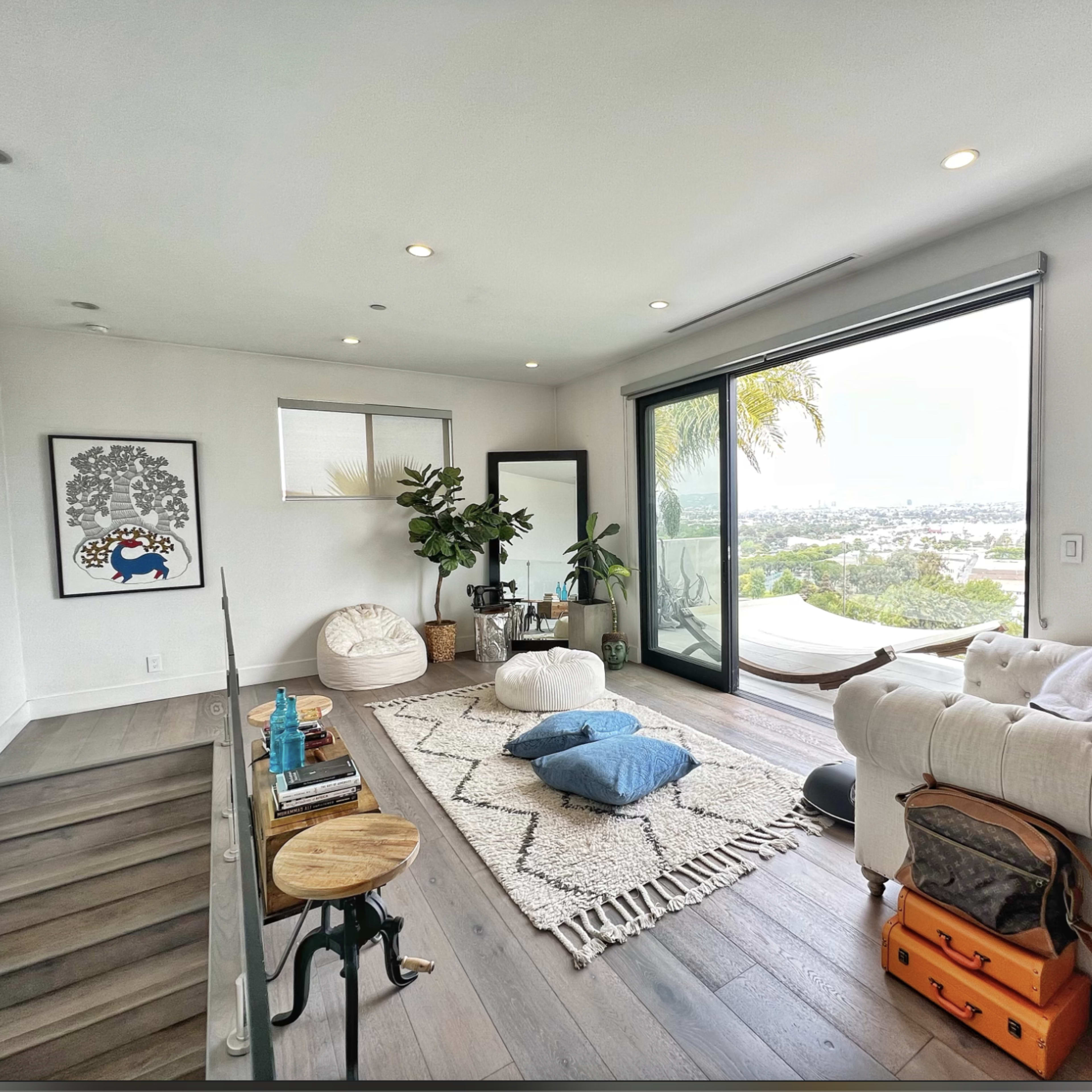 A modern living room features a white couch, oversized cushions, a patterned rug, and large windows overlooking a view.