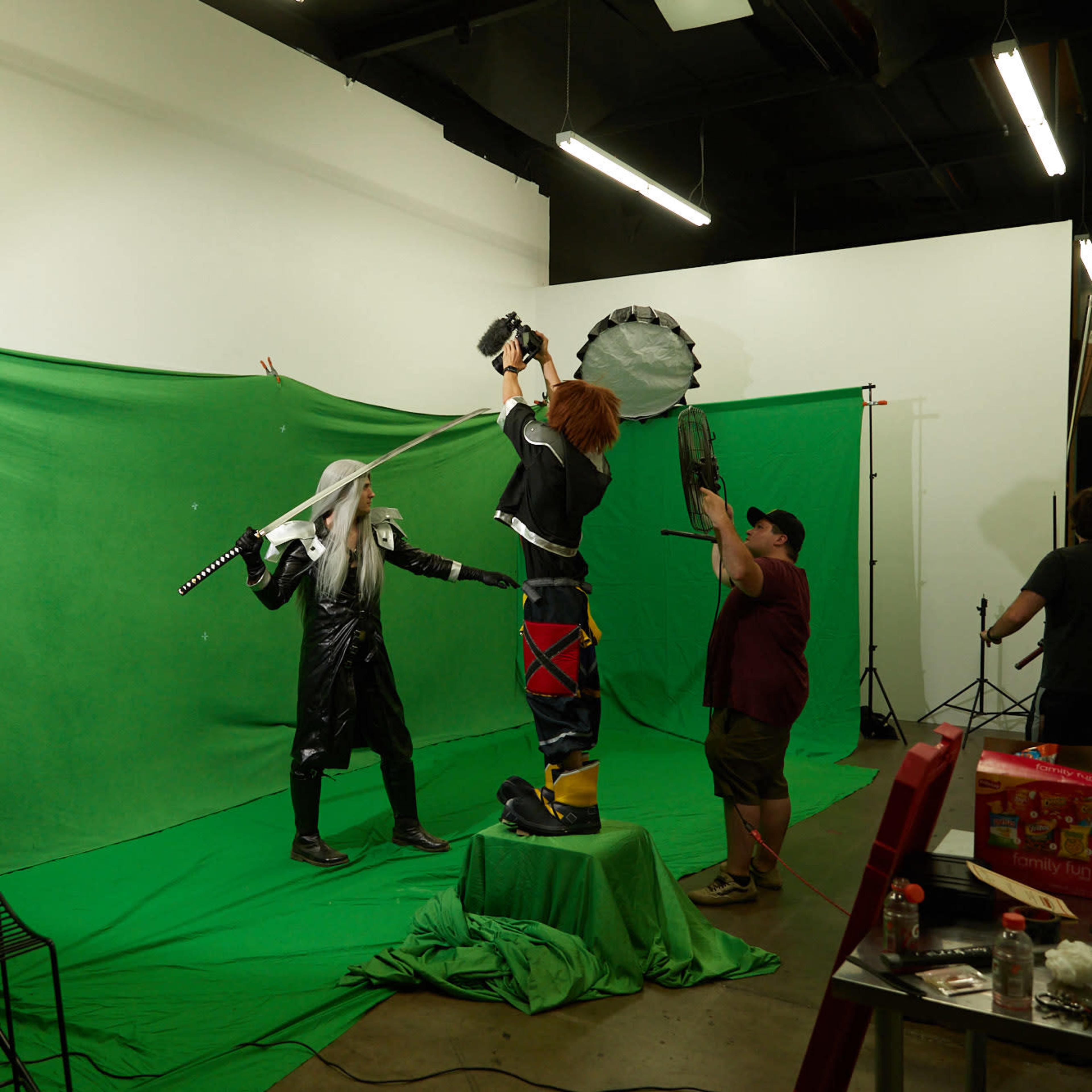 A green screen studio features two individuals in elaborate costumes, one wielding a sword and the other holding an oversized weapon, as crew members prepare for a photoshoot.