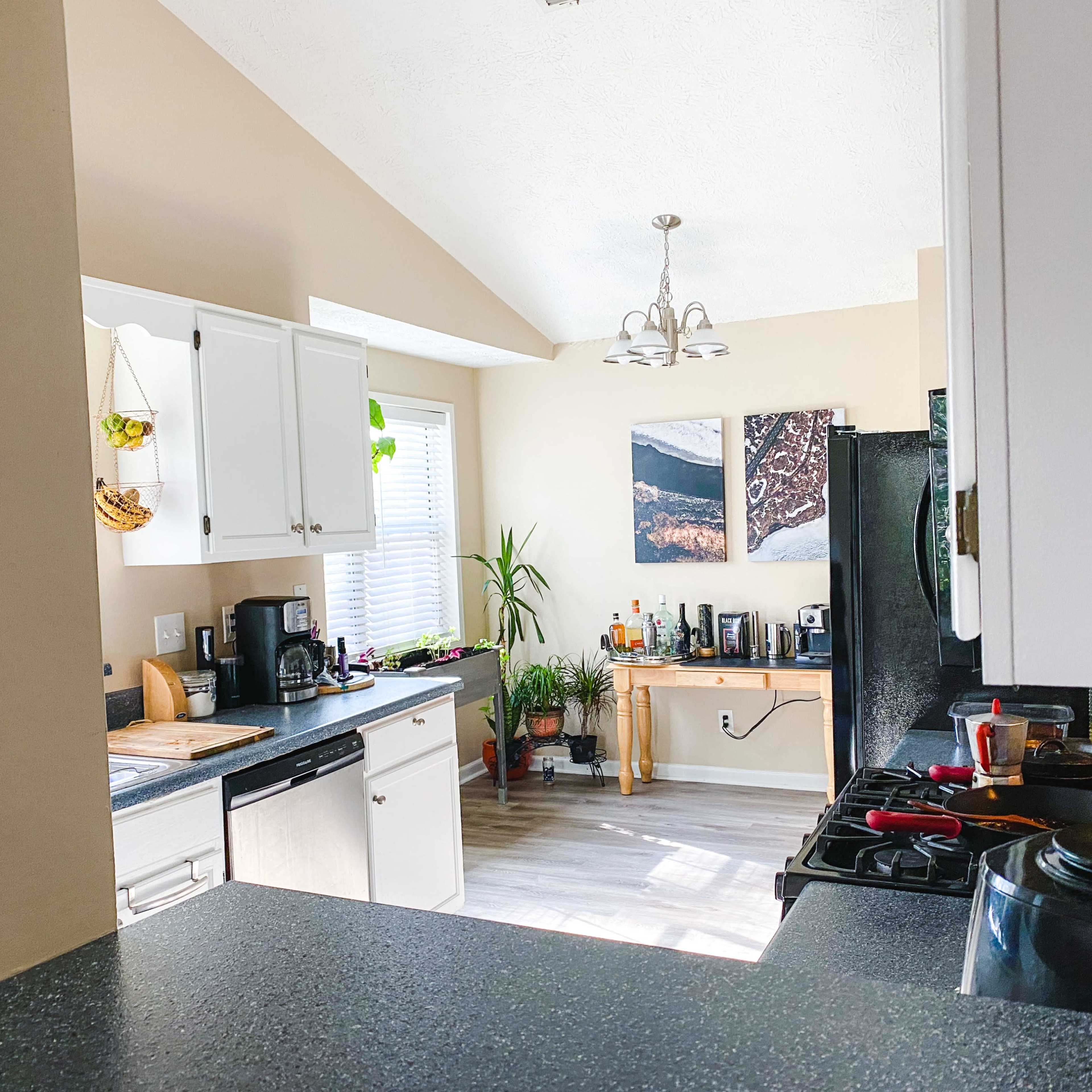 A modern kitchen features white cabinetry, a black countertop, and appliances, with a dining area and plants visible in the background.