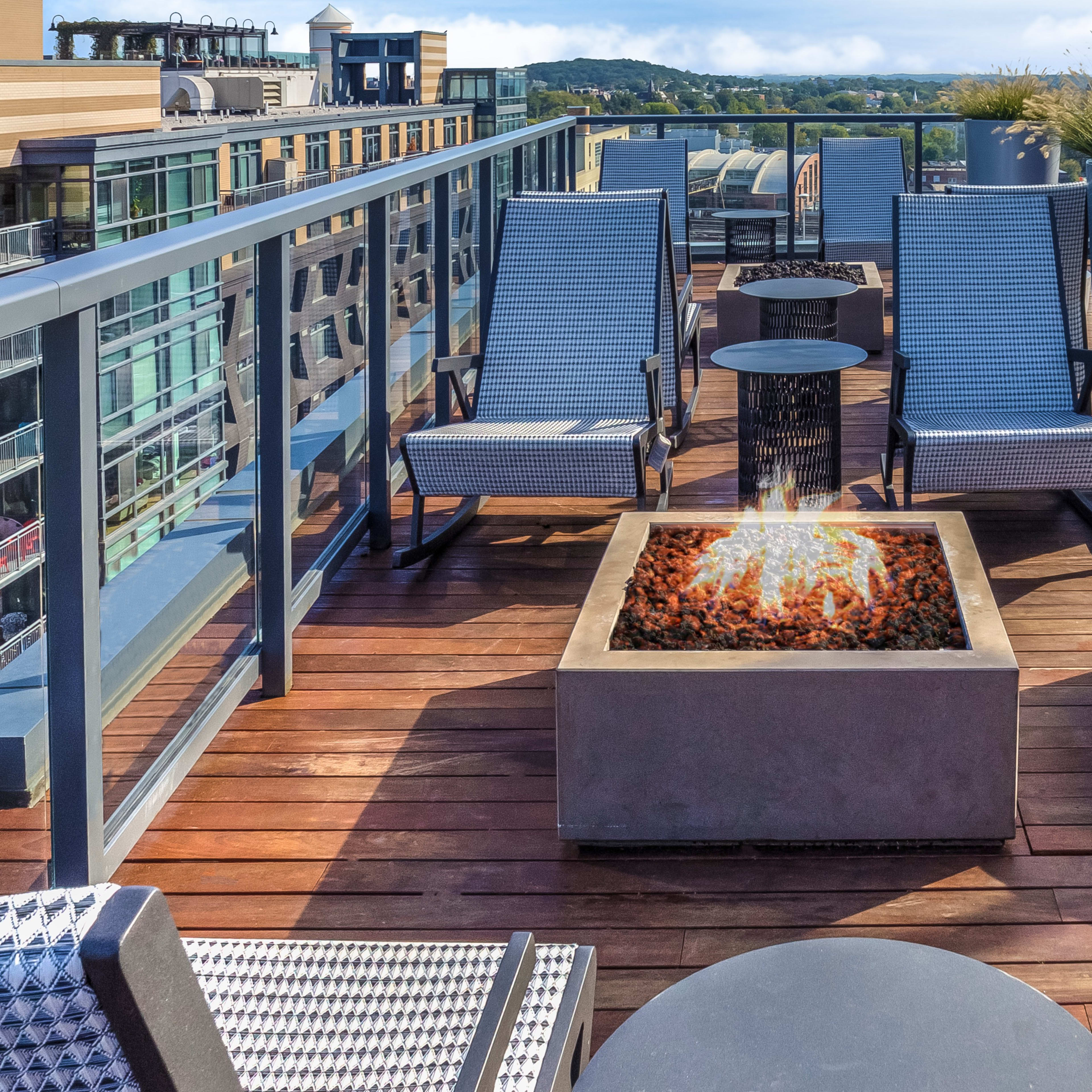 A rooftop terrace with lounge chairs arranged around a fire pit, surrounded by planters and offering a view of the landscape beyond.