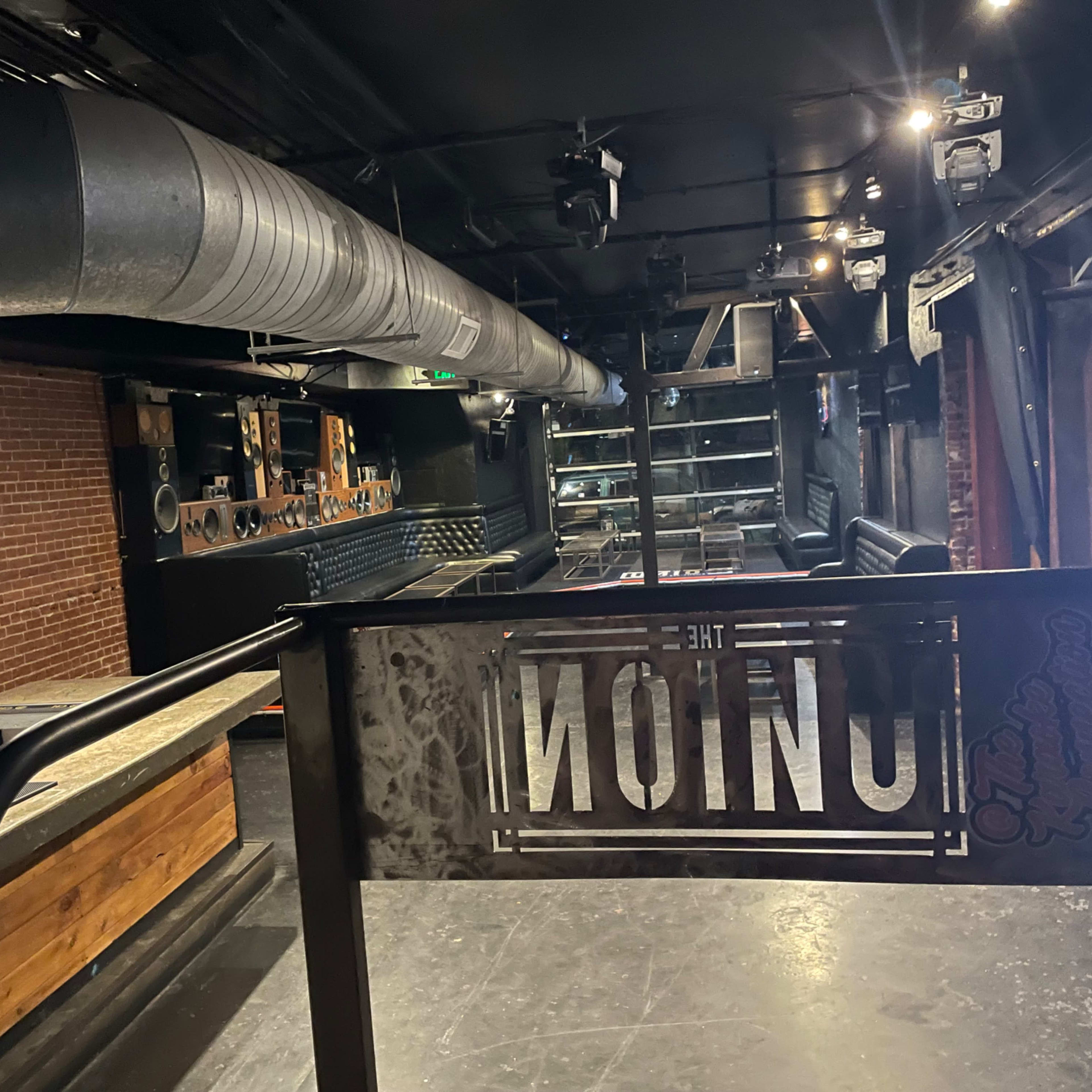 An empty bar interior features a long counter, exposed brick walls, and industrial-style lighting.