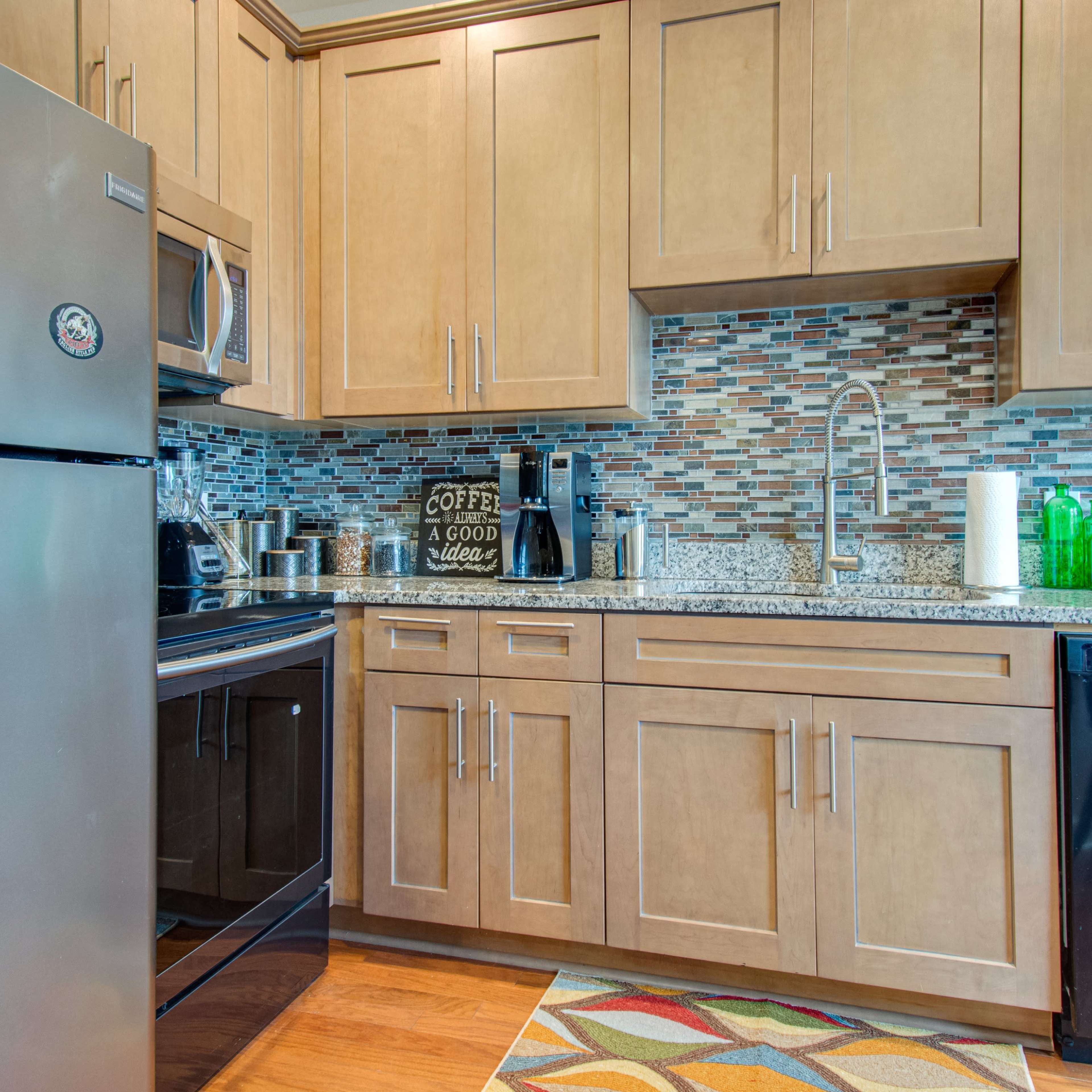 The kitchen features stainless steel appliances, light wood cabinetry, and a mosaic backsplash with a colorful rug on the floor.