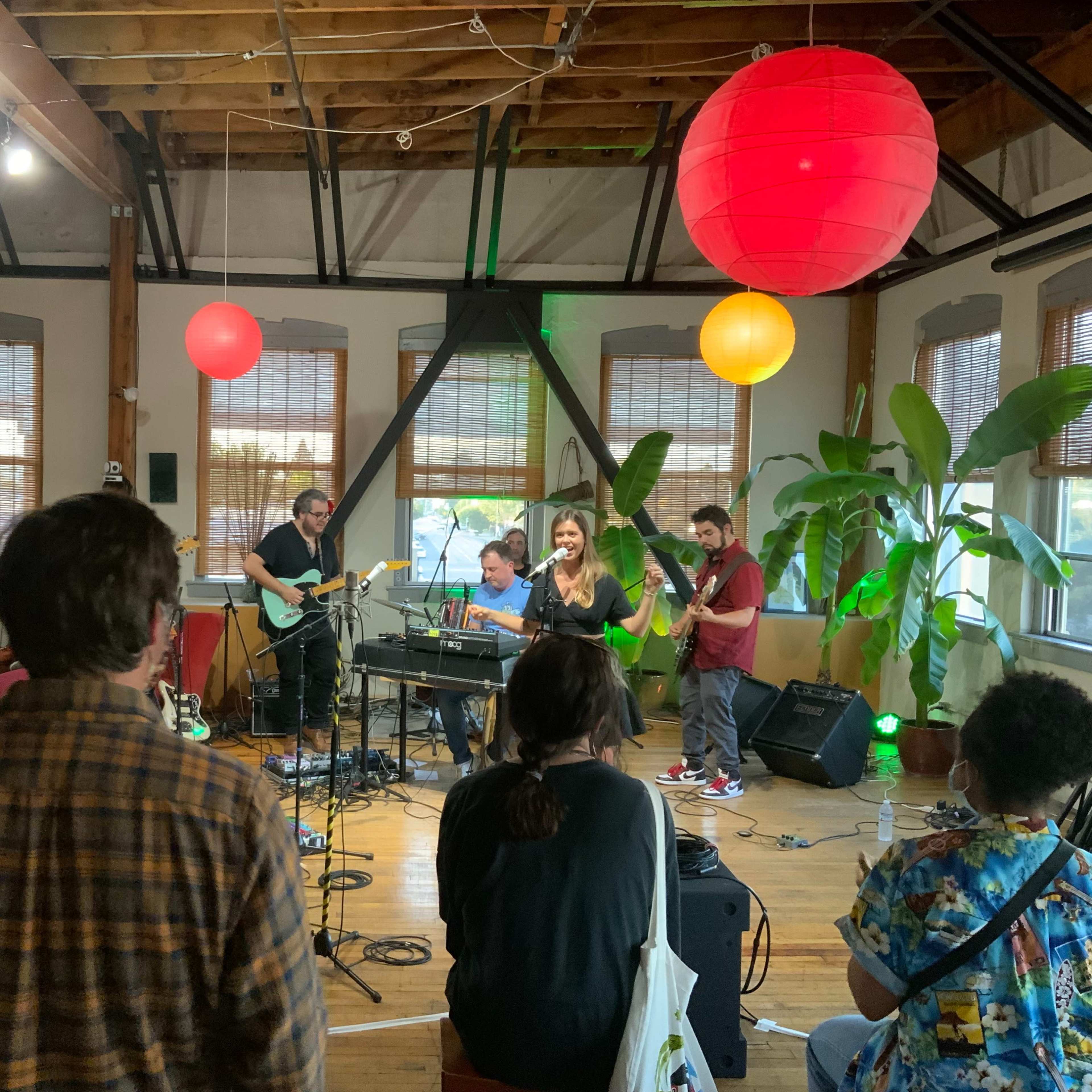 A band performs on stage in a cozy venue with plants and colorful lanterns hanging from the ceiling, while an audience listens attentively.