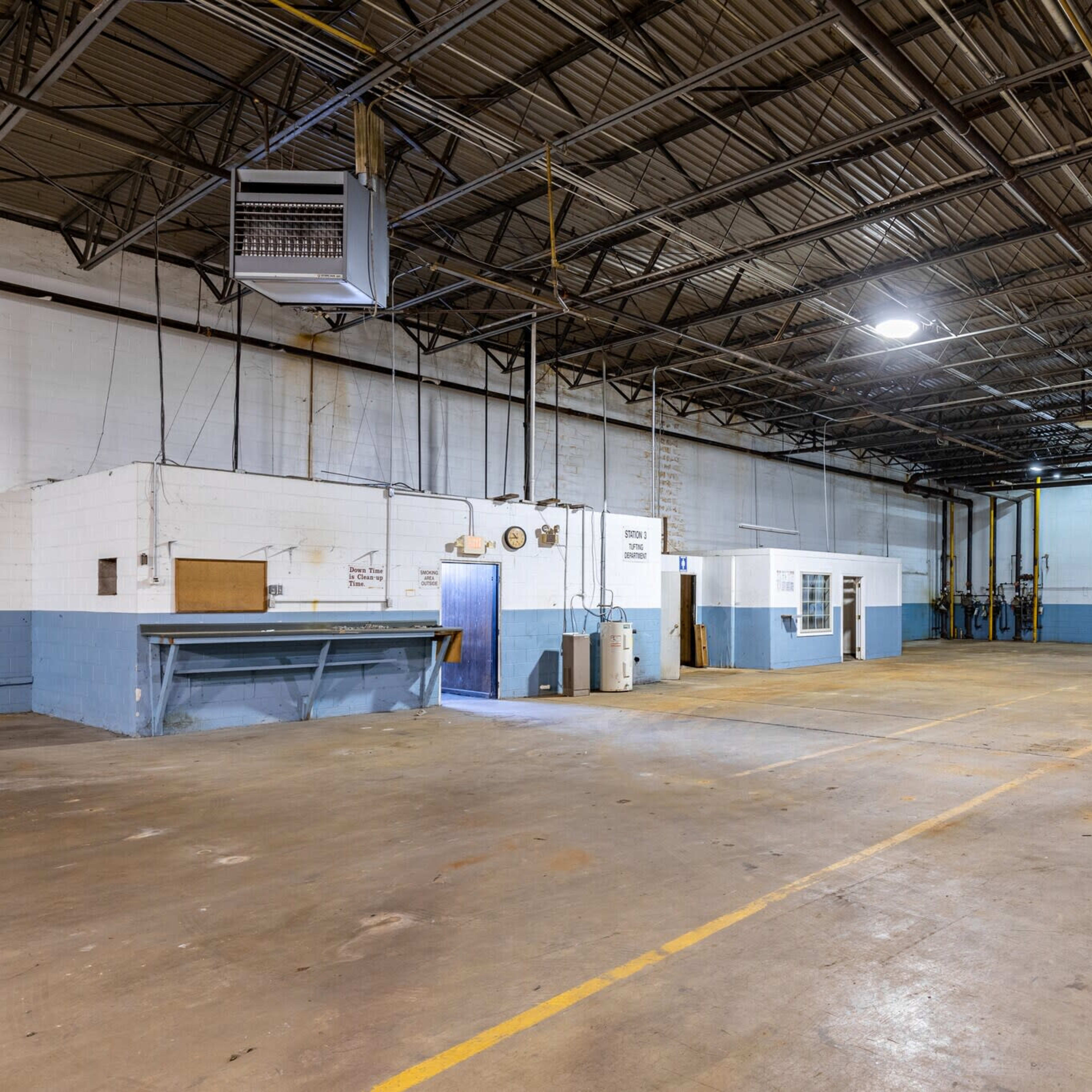 An empty industrial warehouse with a concrete floor and high ceilings, featuring blue-painted walls and overhead lighting.