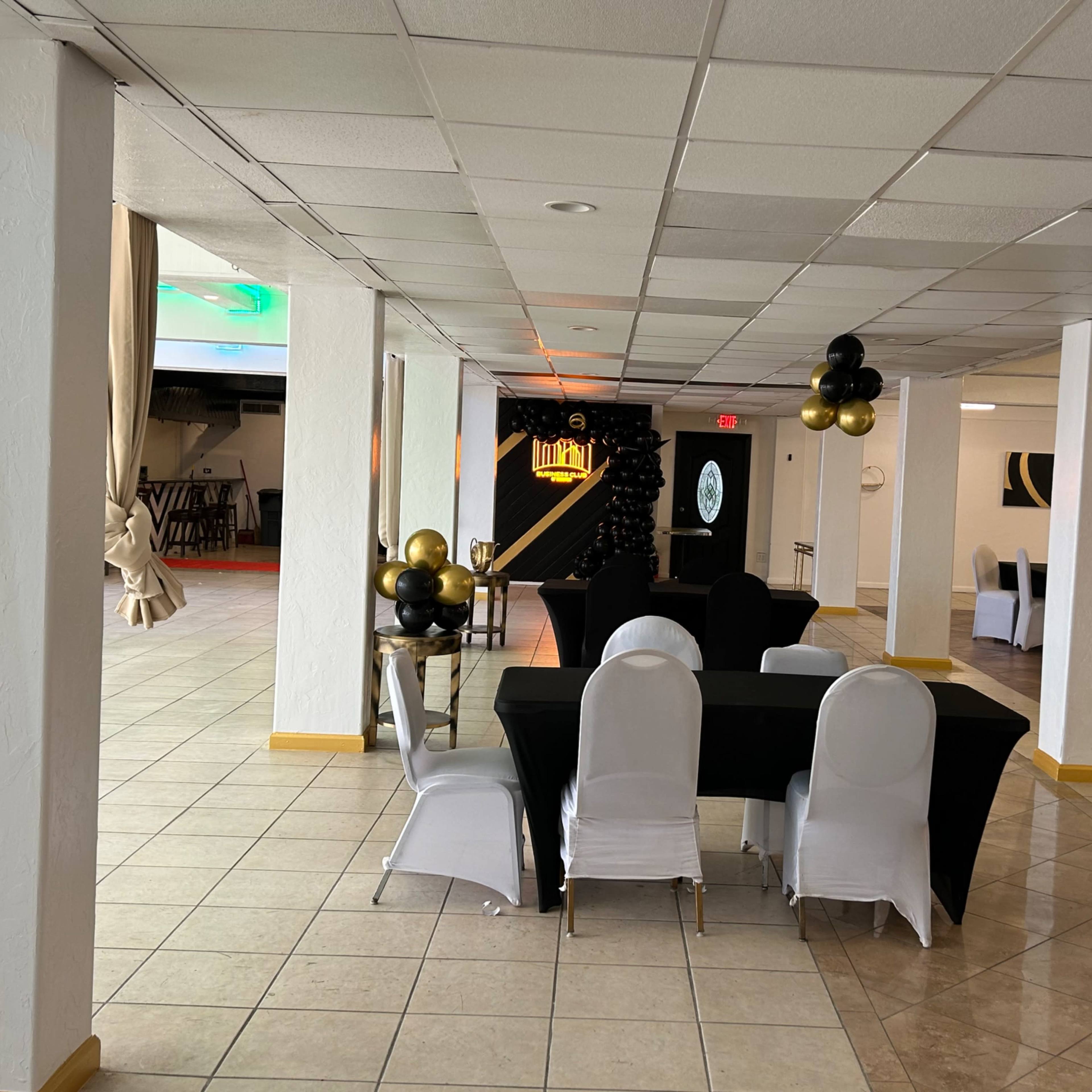 A spacious event venue with tables covered in black and white linens, decorated with balloons, and a staged area at the far end.