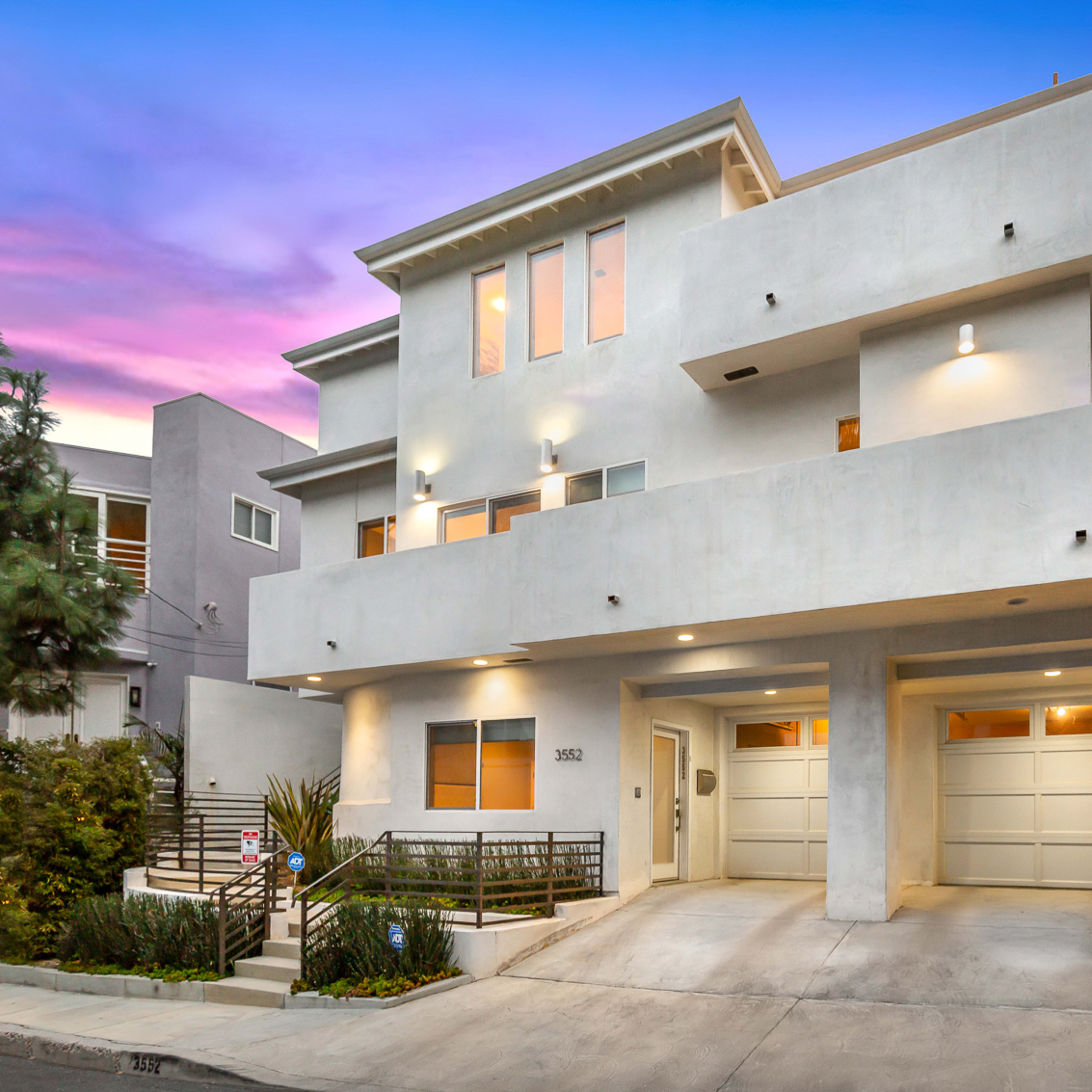 A modern three-story home with a smooth exterior, featuring two garage doors and landscaping, sits at the end of a driveway under a colorful sunset sky.