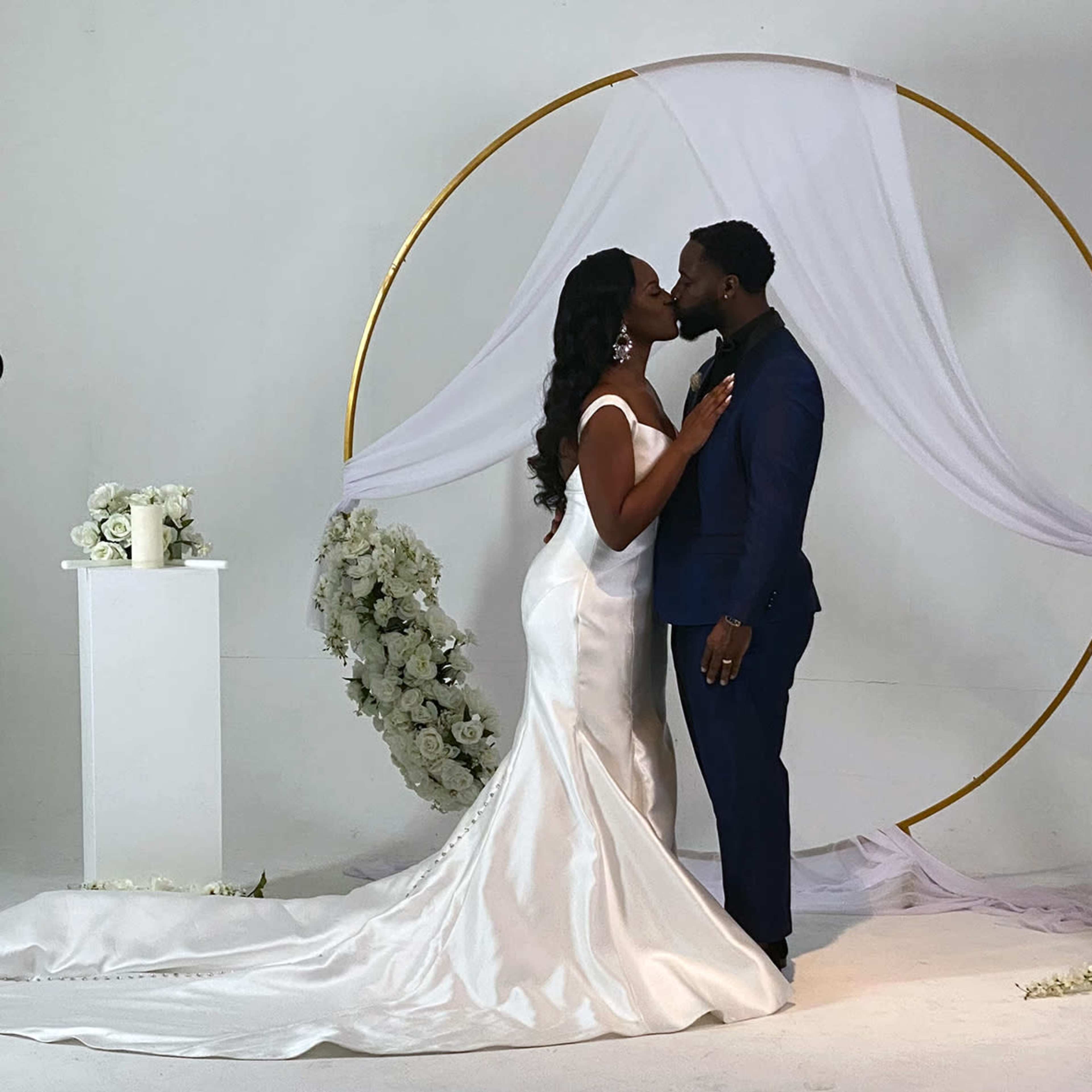 A bride and groom kiss in front of a floral archway draped with white fabric.
