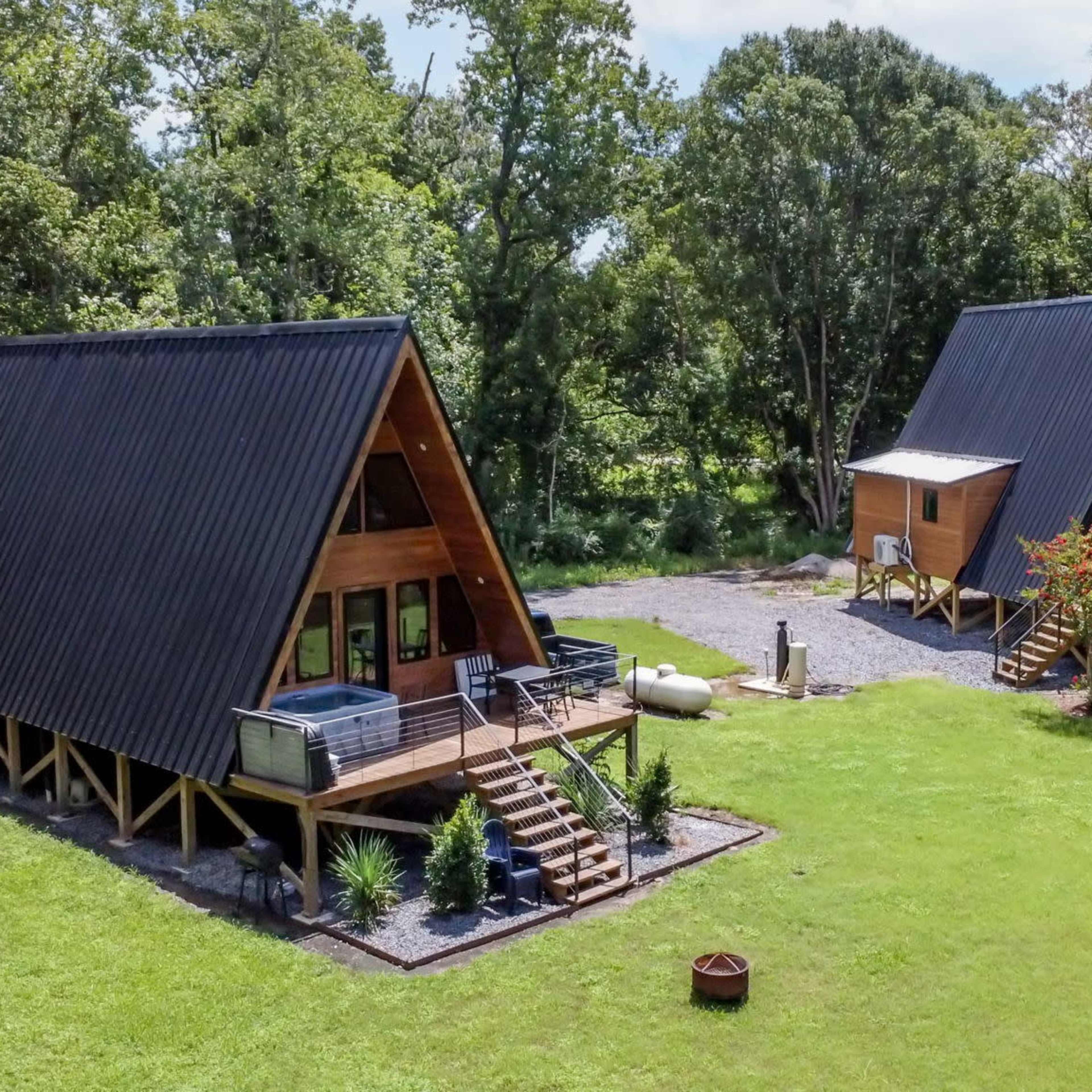 Two A-frame cabins with black roofs are situated on a grassy area surrounded by trees.