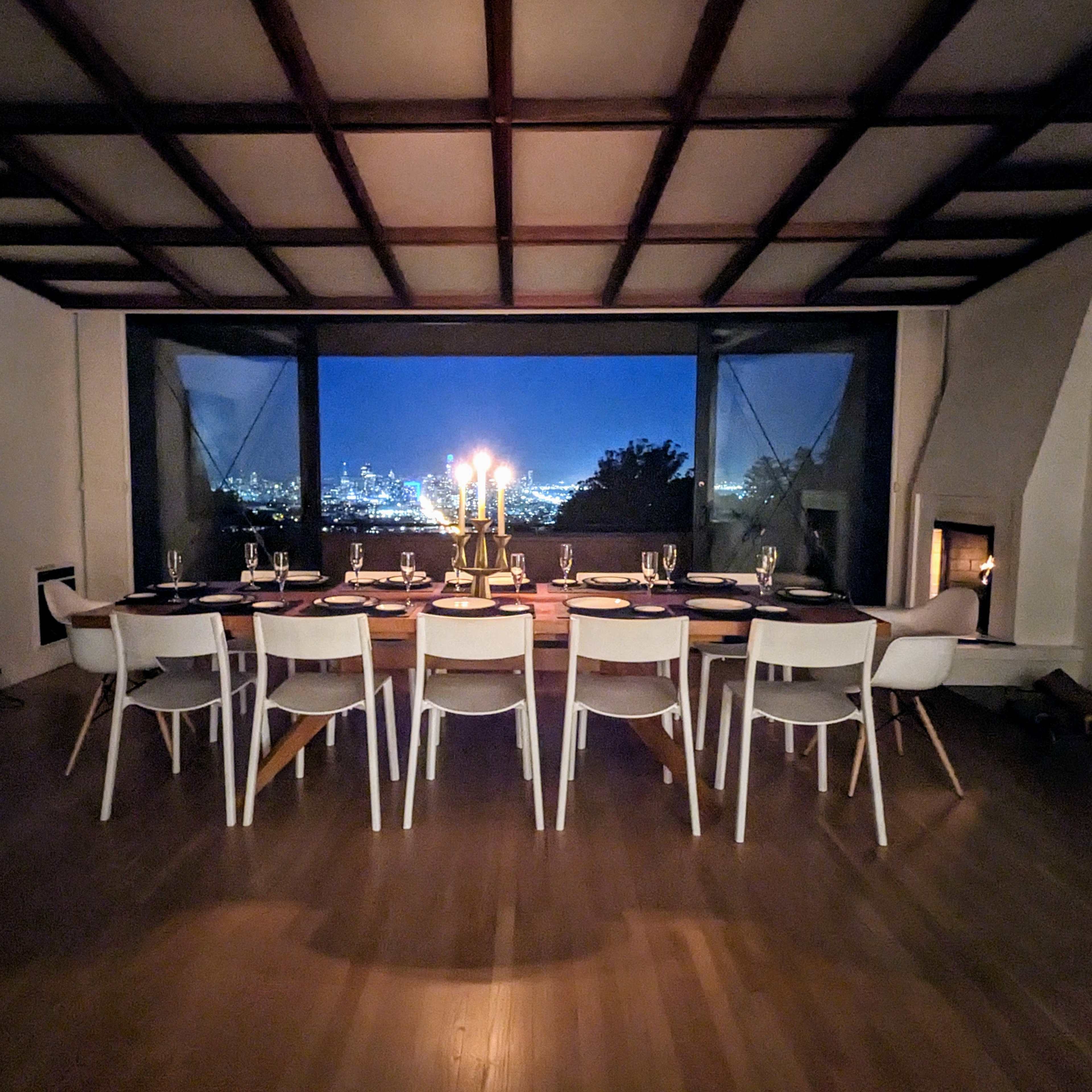 A long dining table is set for a meal, illuminated by candles, with a view of a city skyline at night through large windows.