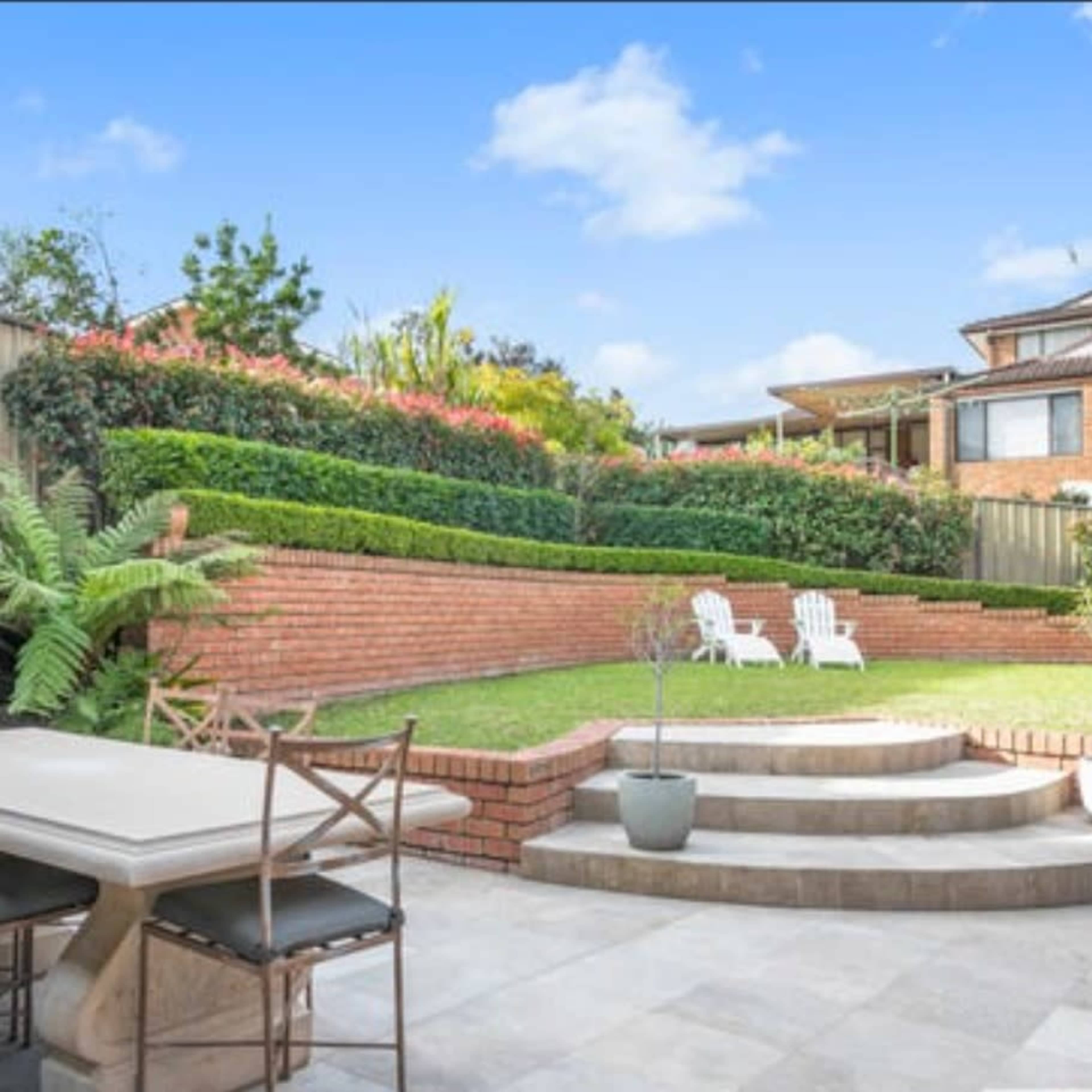 A well-maintained backyard features tiered brick landscaping, two white chairs, and a dining area with a stone table.