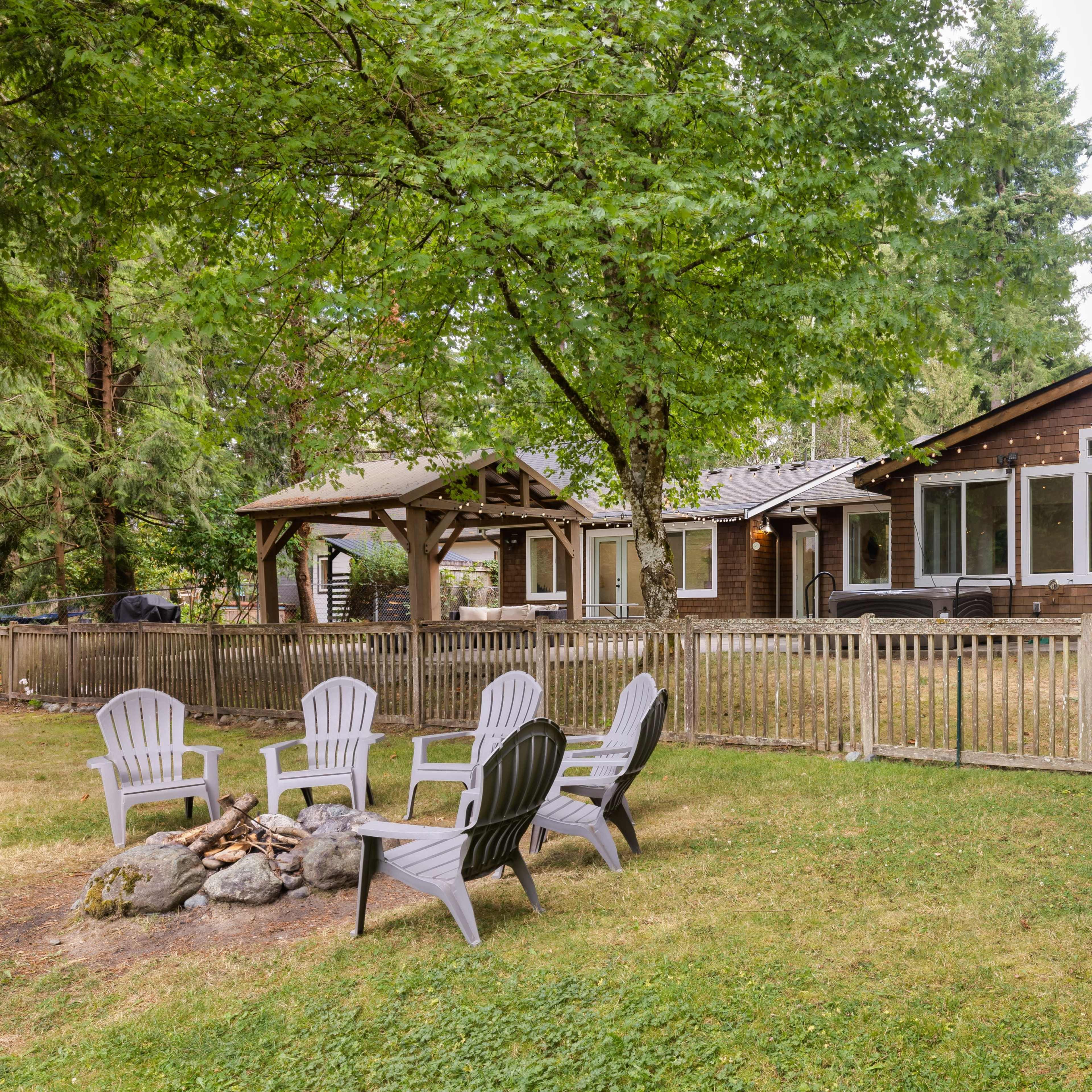 A wooden house with large windows is surrounded by trees and features a fenced yard with a fire pit and several Adirondack chairs.