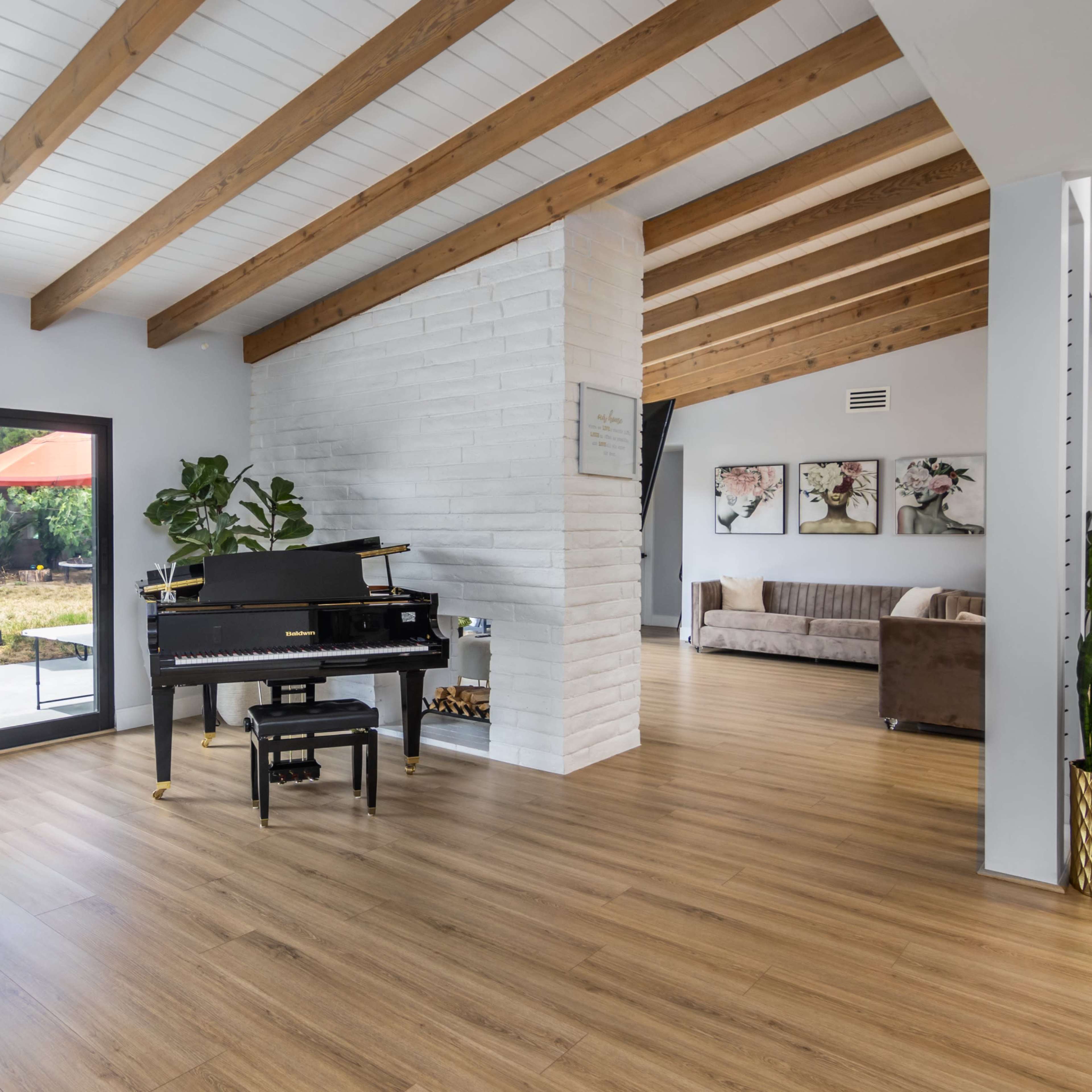 A spacious interior features a black piano positioned near large windows, with a view of a patio and wooden beams overhead.