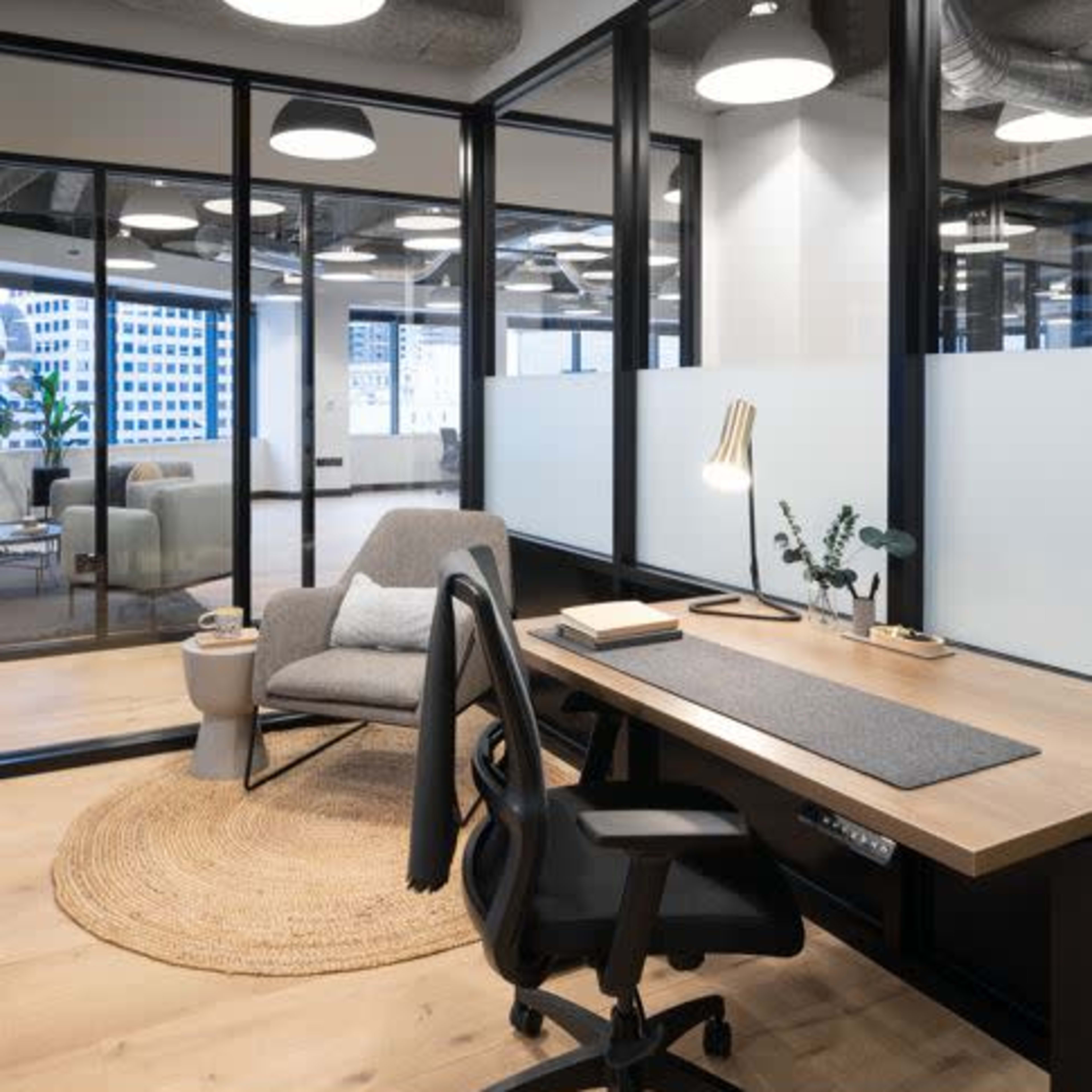 A modern office space featuring a desk with a chair, a cozy armchair, and large glass partitions allowing natural light.