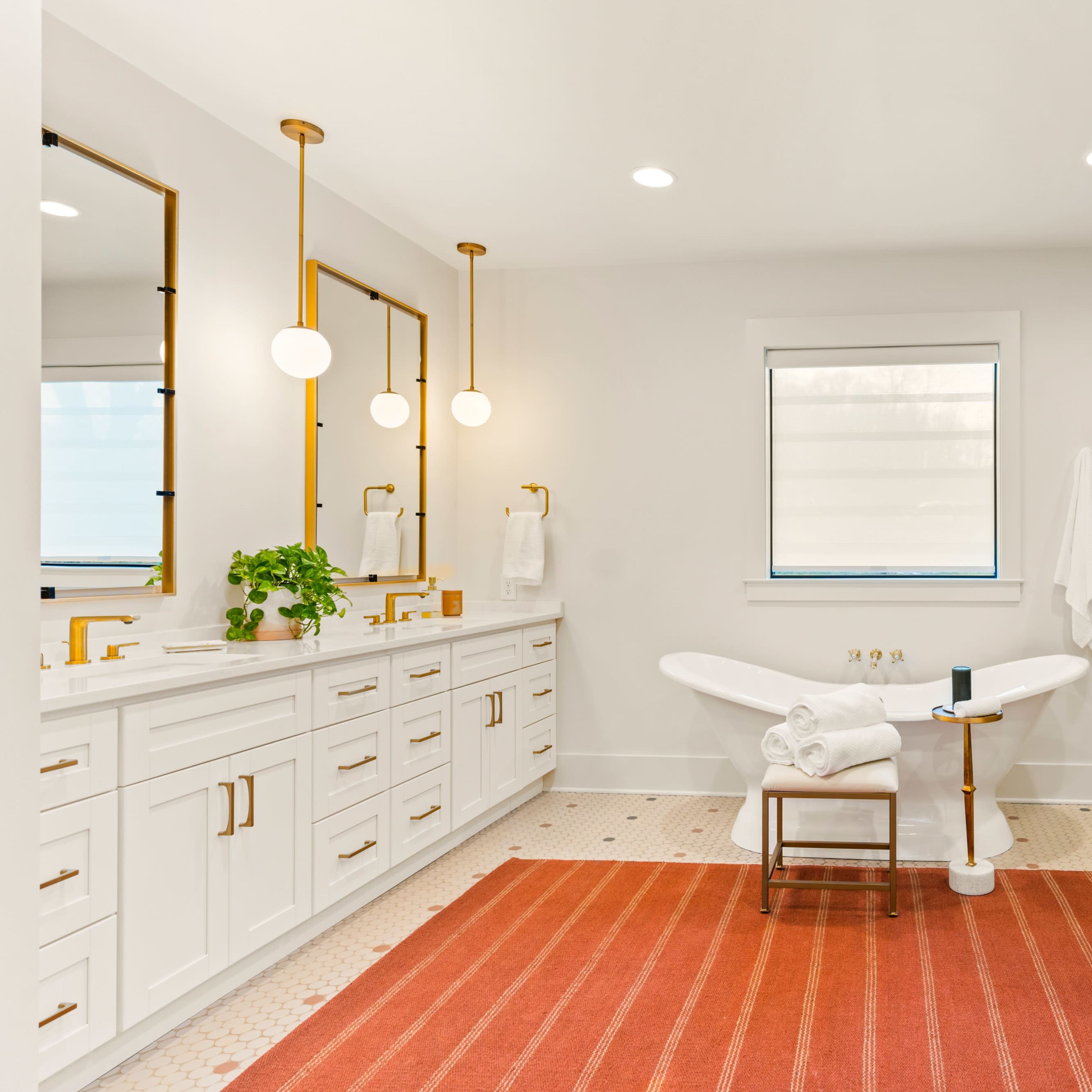 A modern bathroom featuring a freestanding bathtub, a glass shower, double sinks with gold fixtures, and a striped rug on the floor.